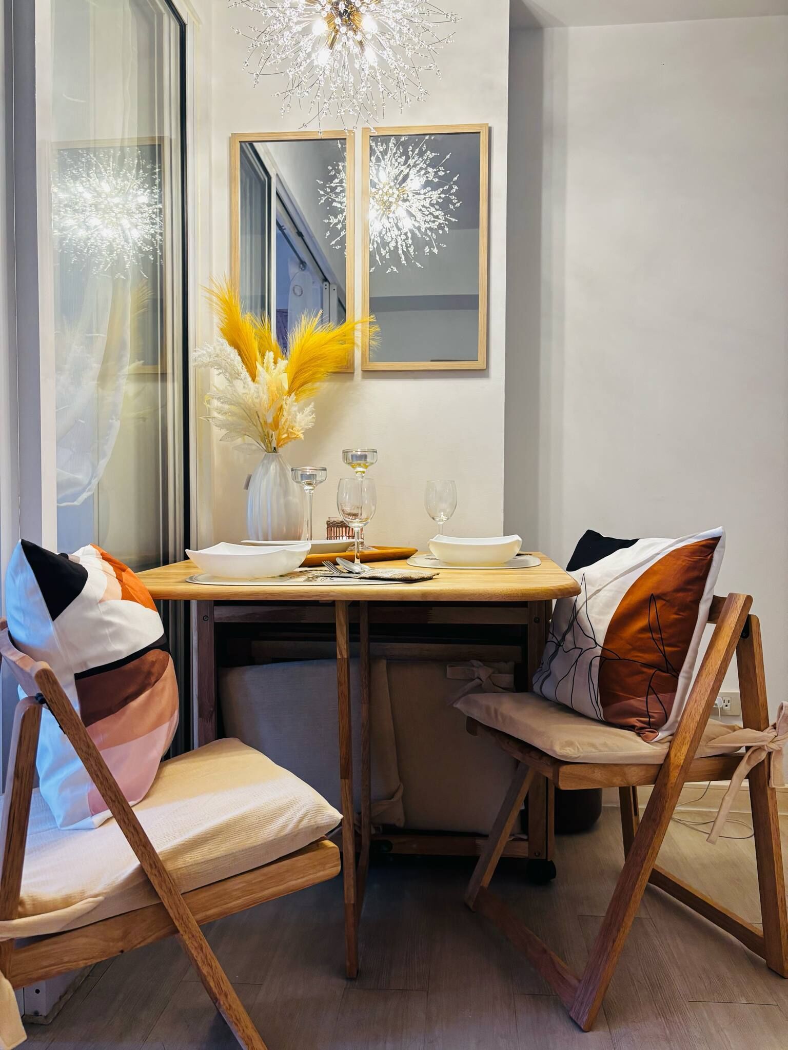 A dining area with a wooden table, two chairs, a vase with dried flowers, and a mirror on the wall.