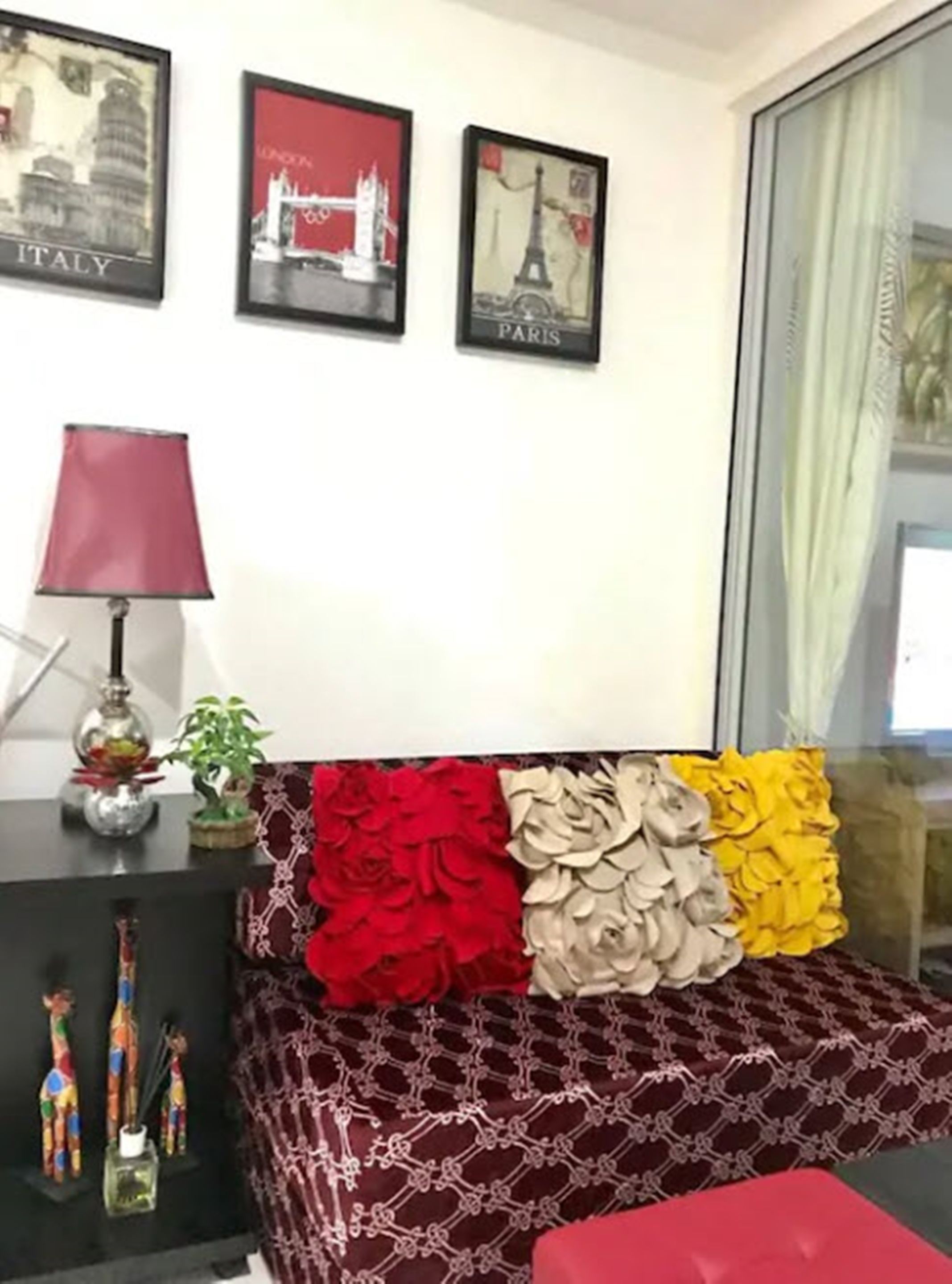 A living room with a sofa adorned with red, beige, and yellow pillows, a red lamp, and framed pictures of Italy and Paris on the wall.