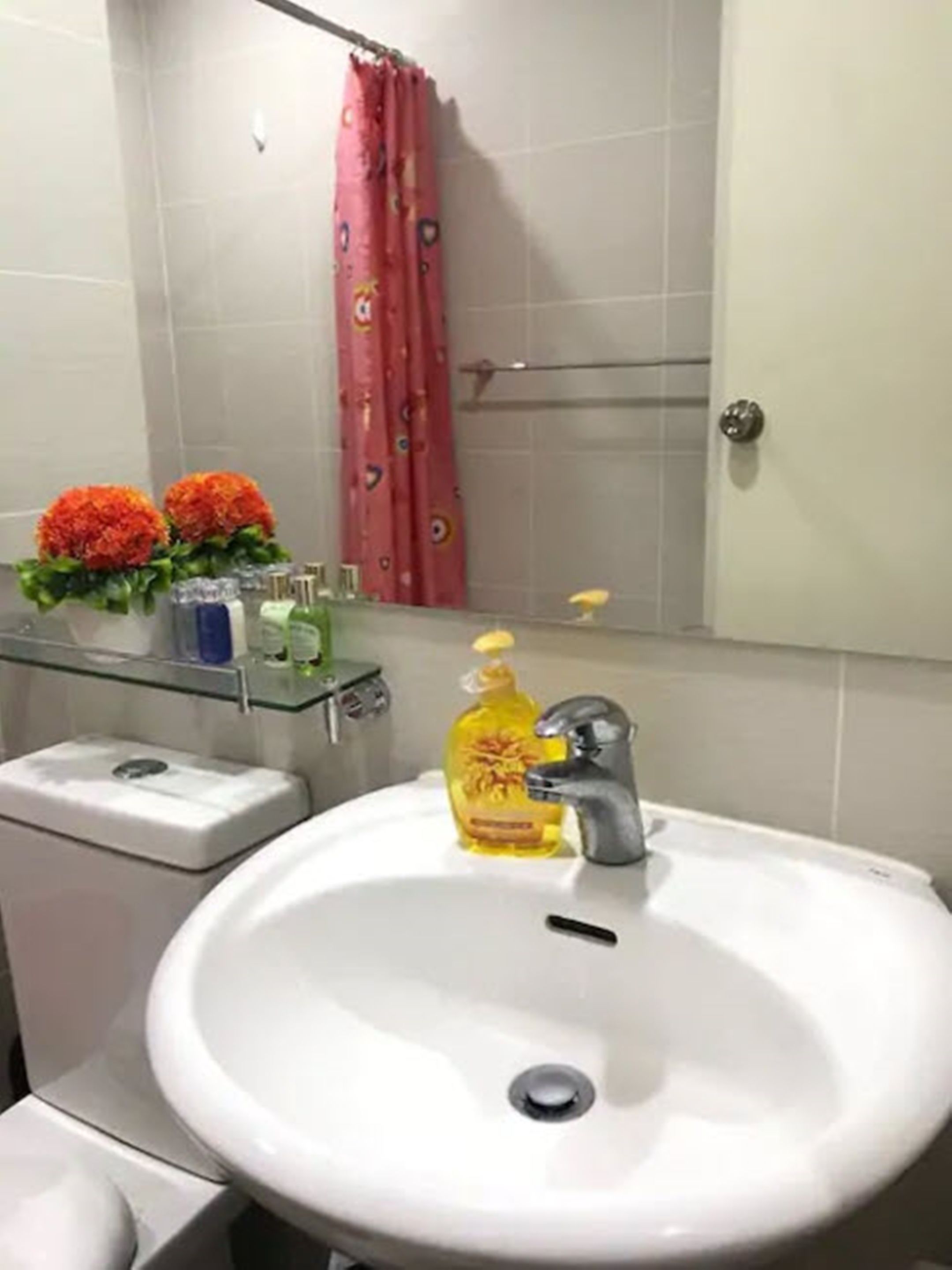 Un baño con lavamanos blanco, inodoro, repisa de vidrio con artículos de aseo y cortina de ducha.