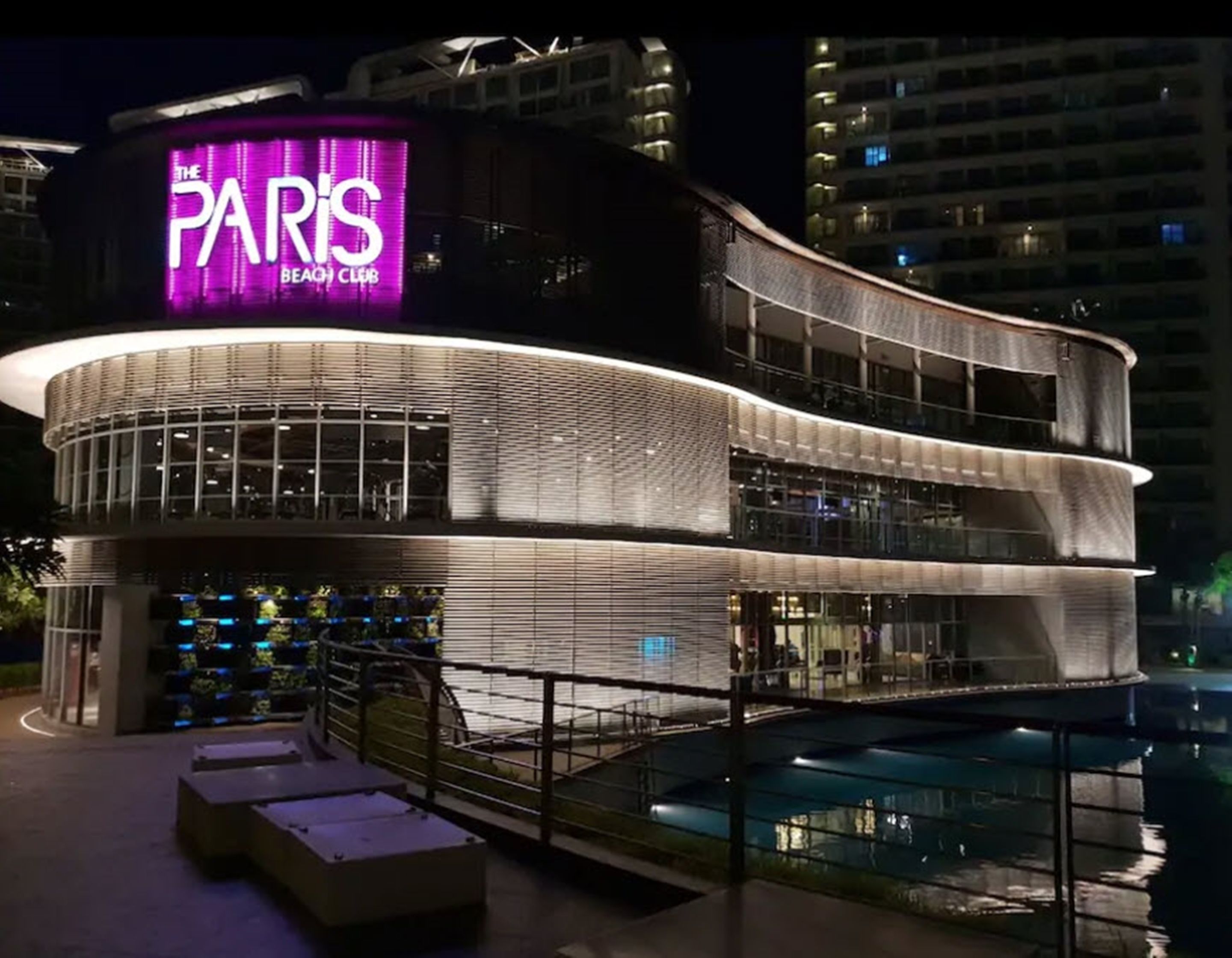 A modern building with a prominent sign reading "The Paris Beach Club." It features a curved facade with multiple balconies and a pool area visible in the foreground.