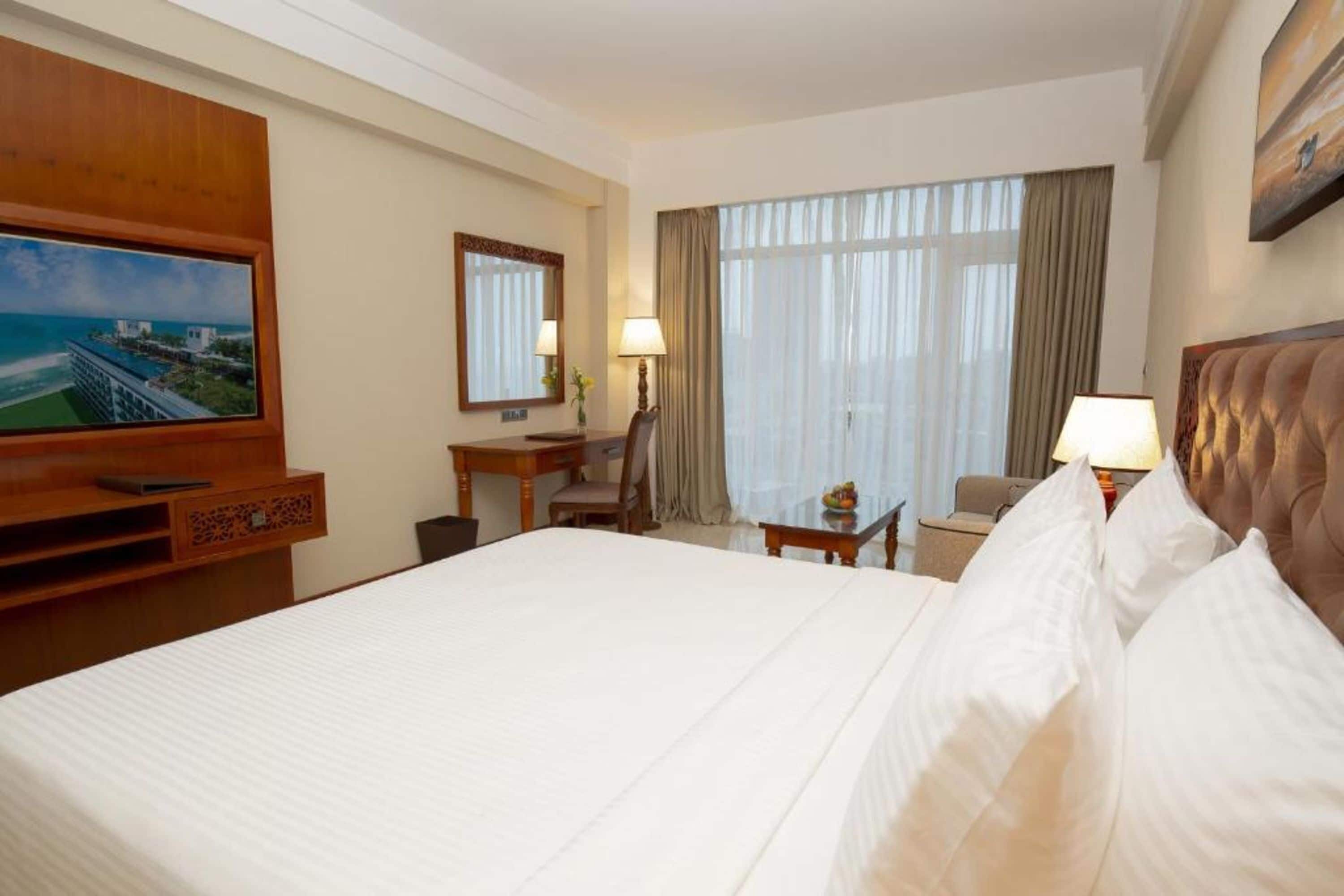 Quarto de hotel com uma cama grande, uma escrivaninha com cadeira, uma TV na parede e uma janela com cortinas.
