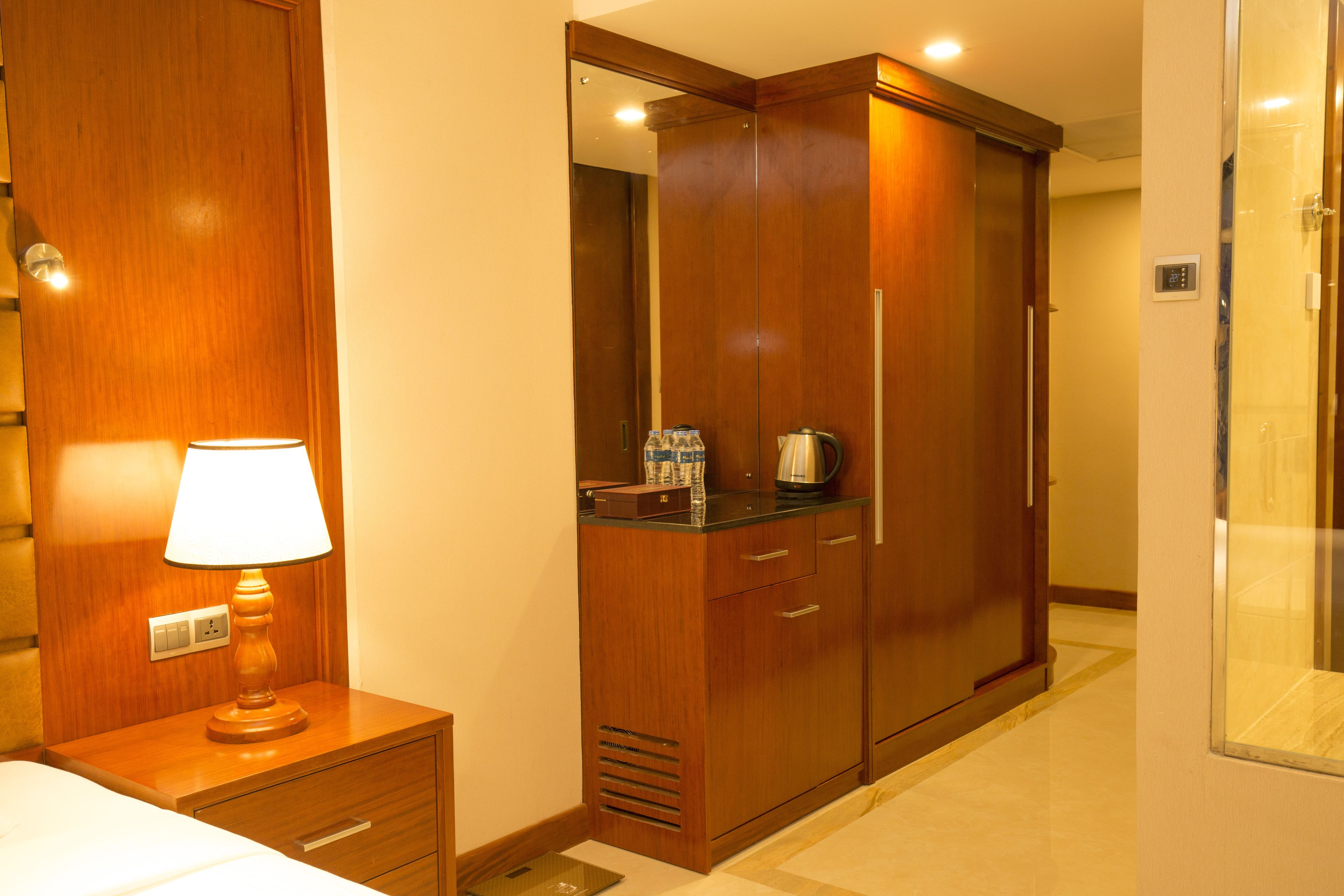 A hotel room with a wooden wardrobe, a bedside table with a lamp, and a small kitchenette area.