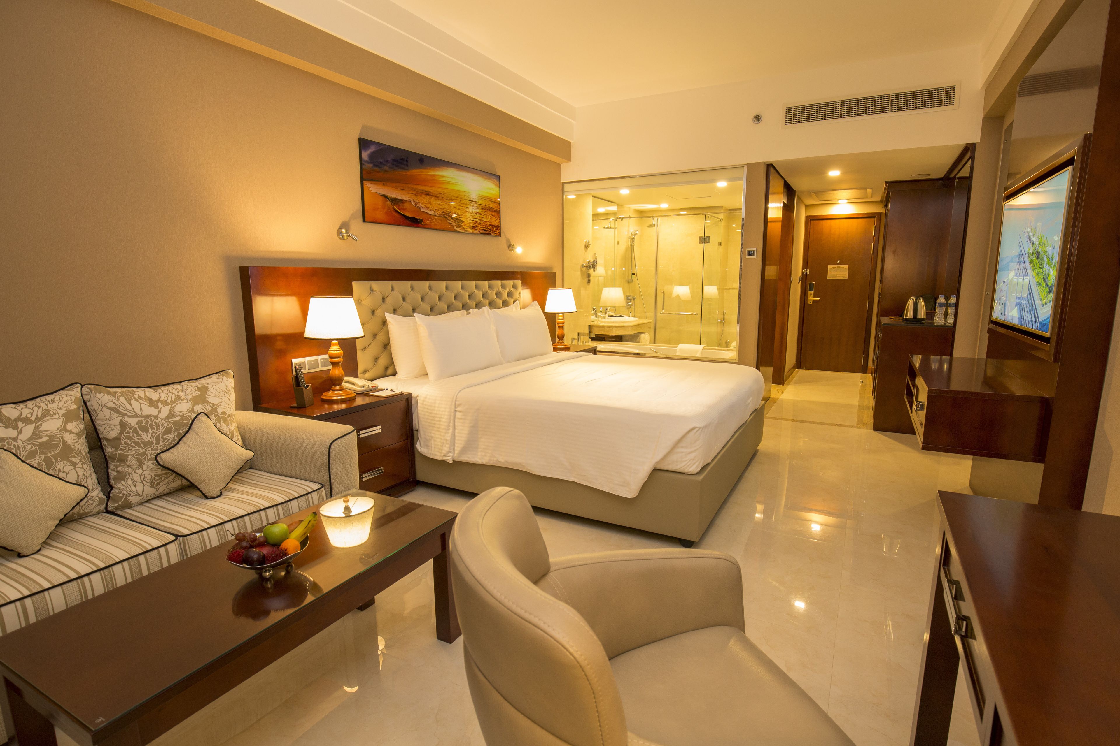 A hotel room with a bed, a sofa, a desk with a fruit bowl, a chair, a nightstand with lamps, and a bathroom visible through a glass door.