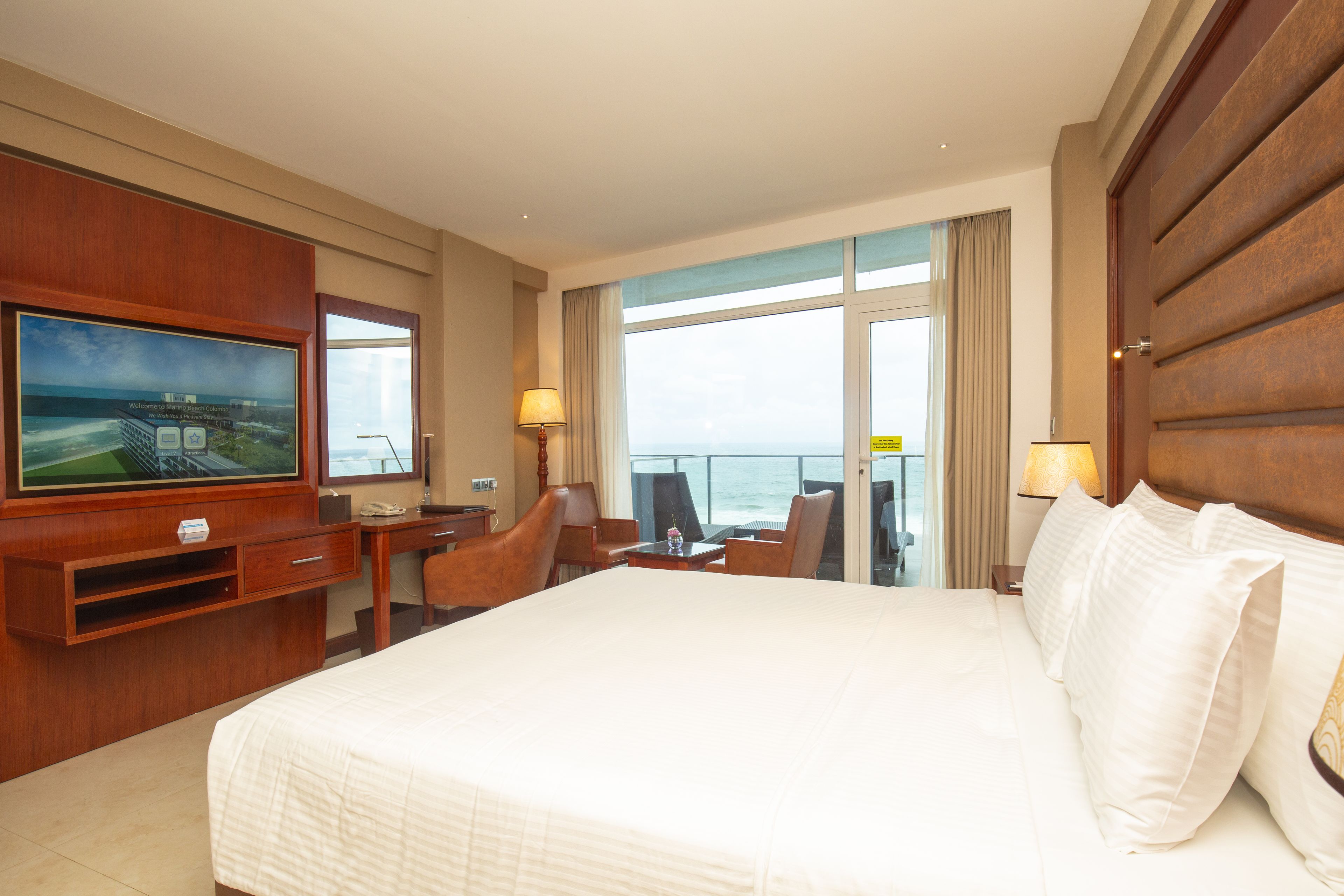 A hotel room with a large bed, a television mounted on the wall, a desk with a chair, and a view of the ocean through the window.