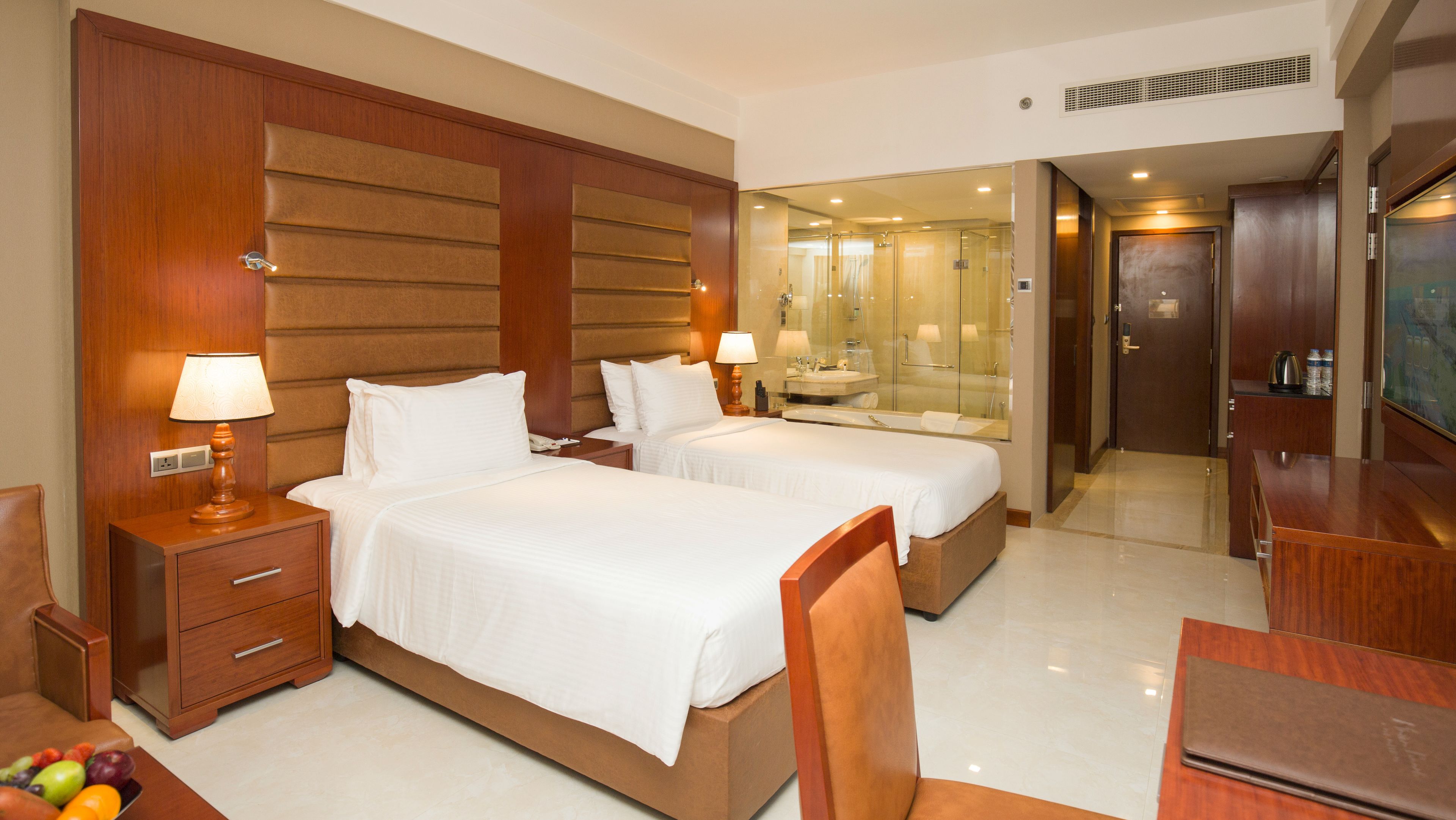 A hotel room with two beds, a desk, a chair, a nightstand, a lamp, a mirror, and a bathroom with a sink and mirror.