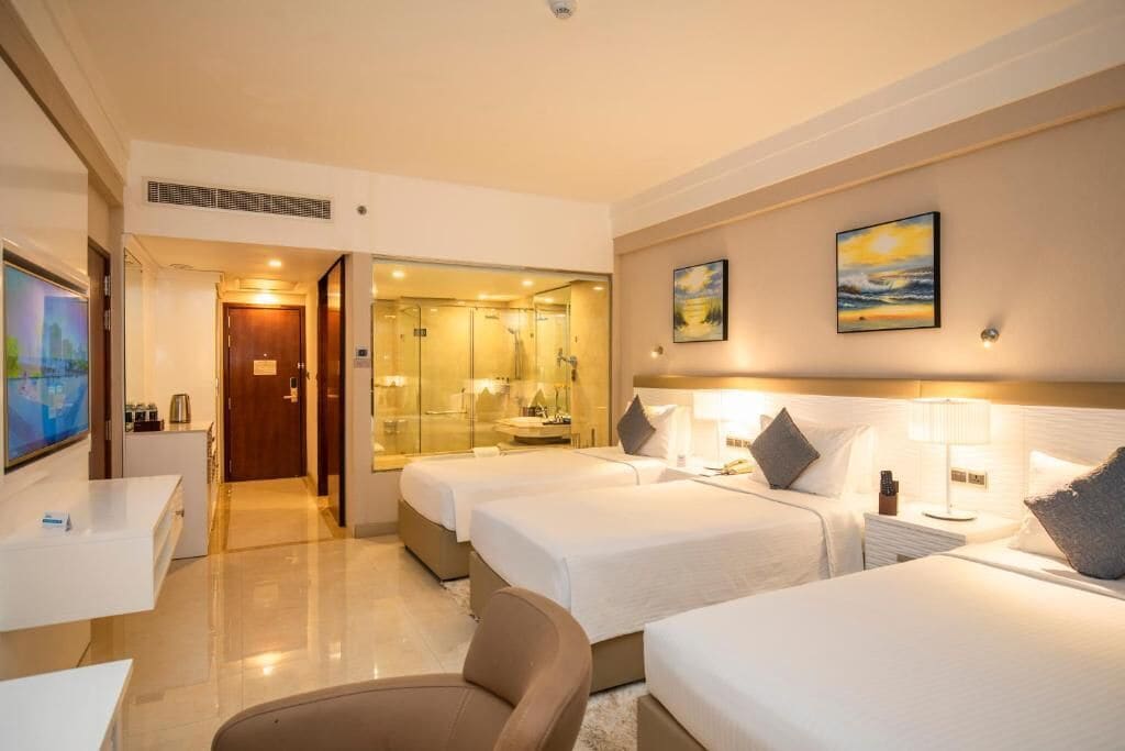 A hotel room with two beds, a desk, a TV, a bathroom with a glass door, and artwork on the walls.
