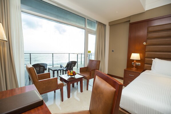 A hotel room with a bed, a desk, two chairs, a small table with fruit, and a view of the sea through large windows.