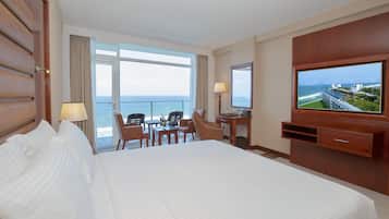 A hotel room with a large bed, a desk with a chair, a television, and a view of the ocean.