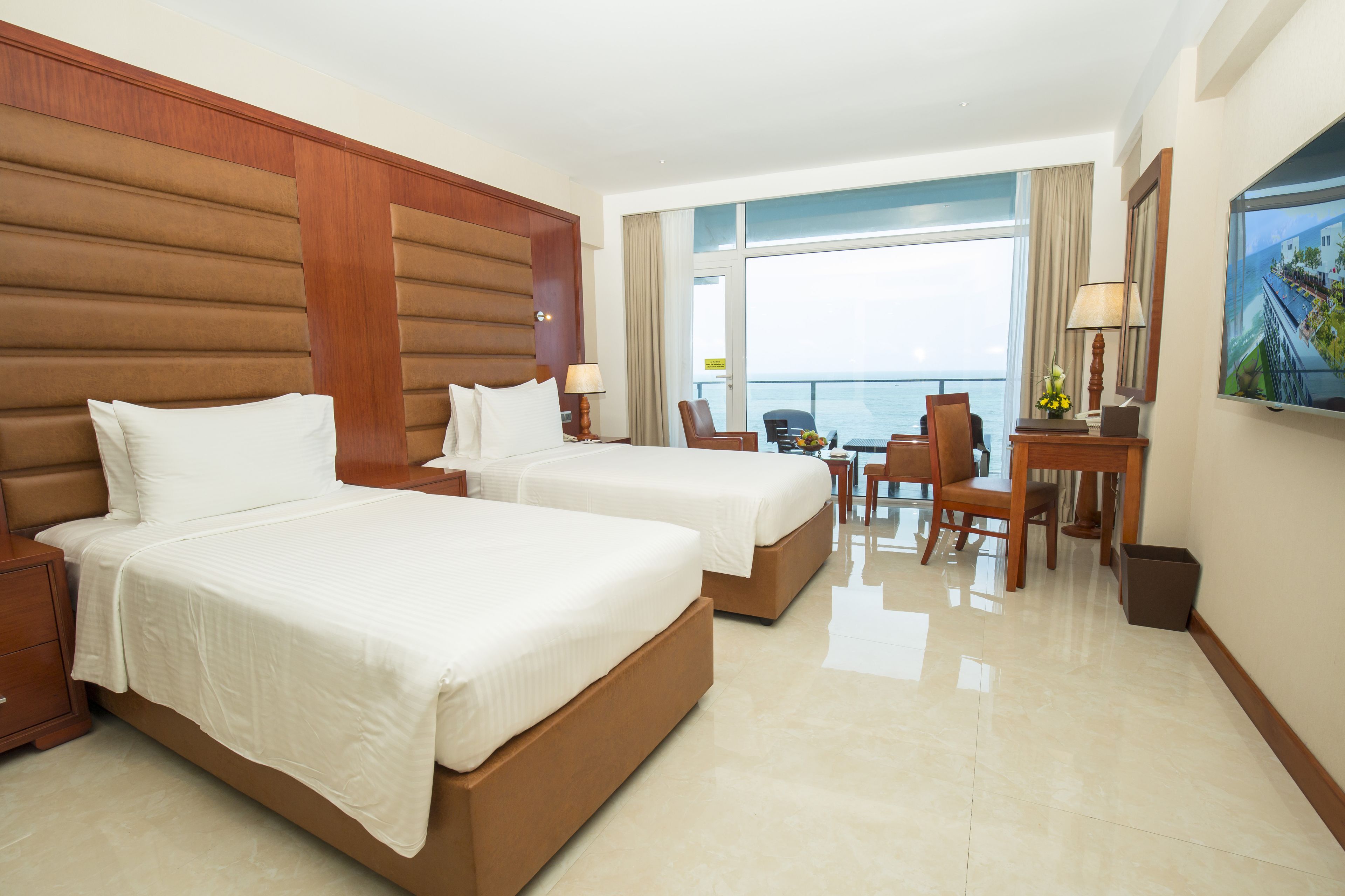 A hotel room with two beds, a desk, chairs, and a large window with a view of the sea.