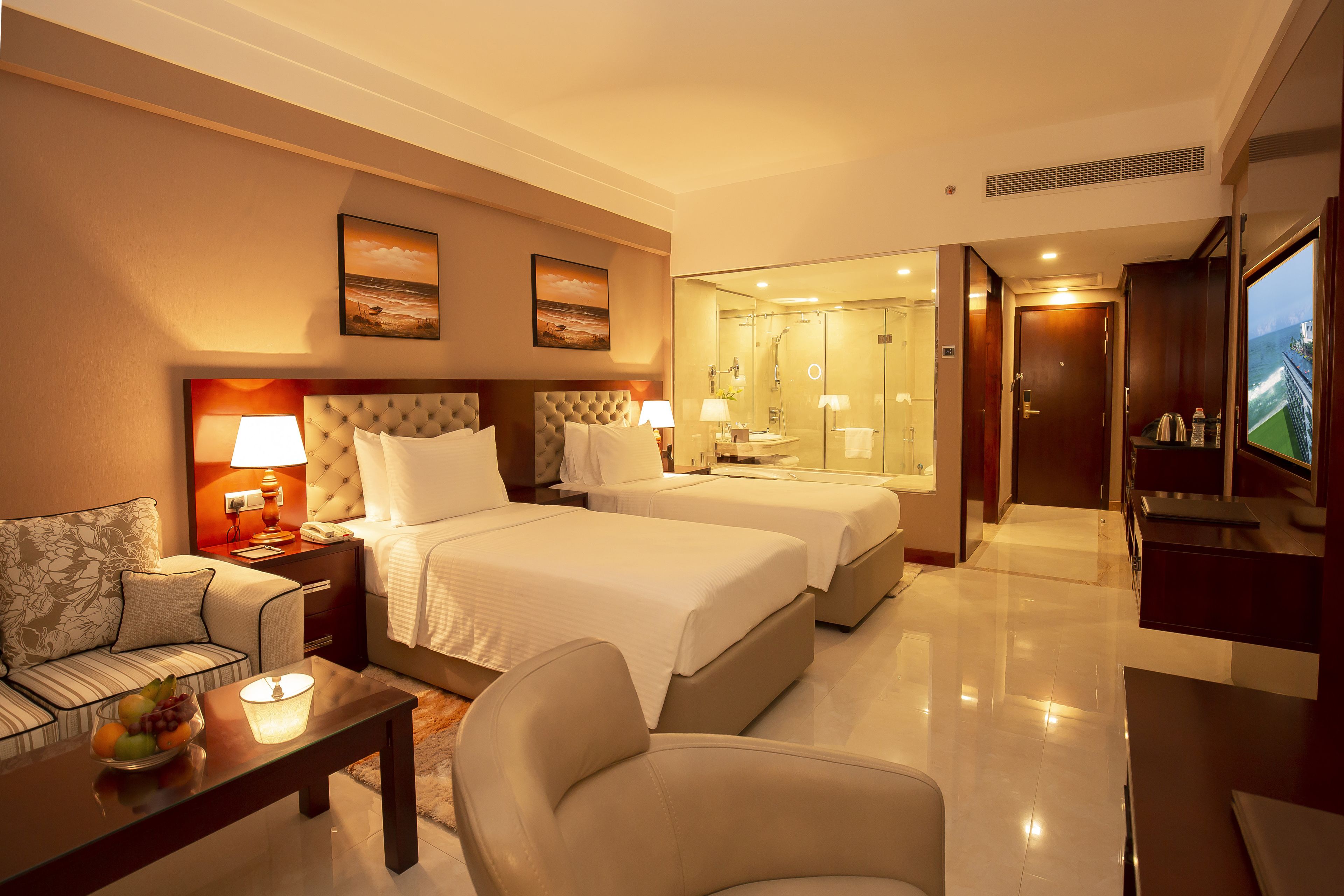 A hotel room with two beds, a sofa, a coffee table with fruit, a TV, and a bathroom visible through a glass door.