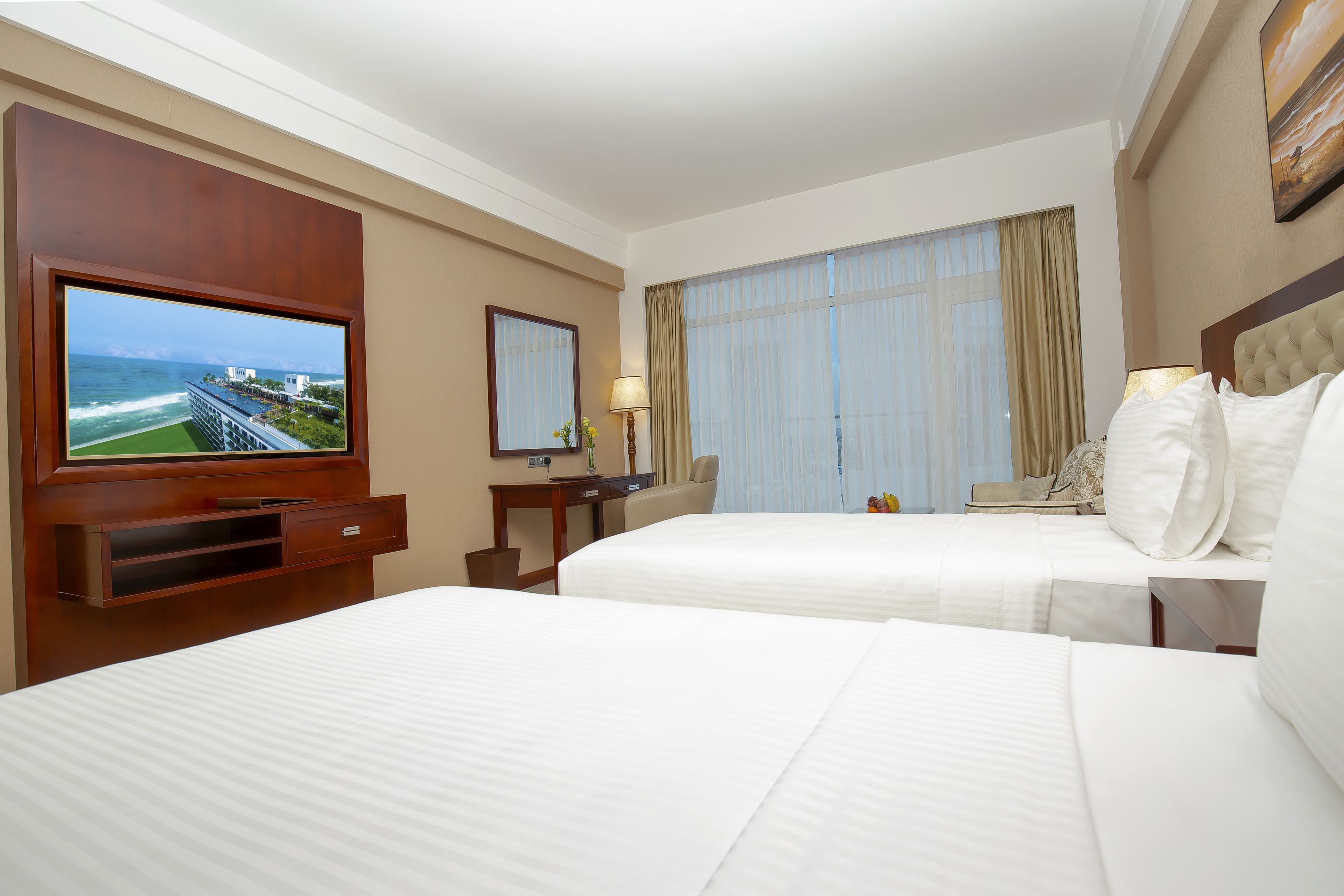 A hotel room with two beds, a TV, a desk, and a large window with curtains.