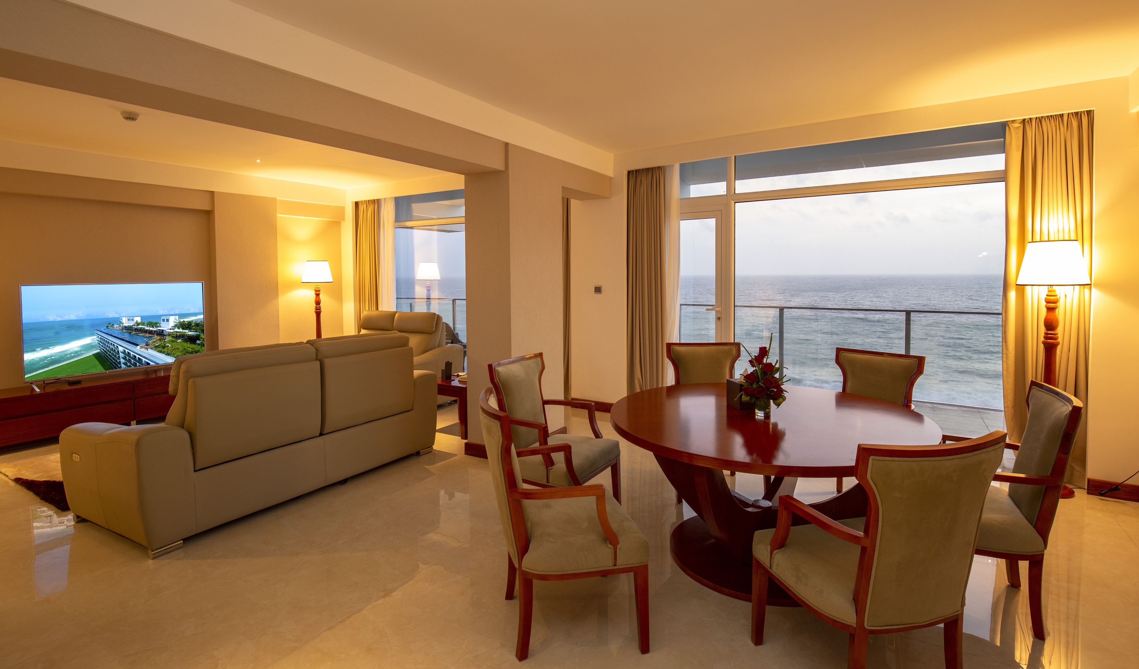 A hotel room with a large TV, a sofa, a dining table with chairs, and a view of the ocean.