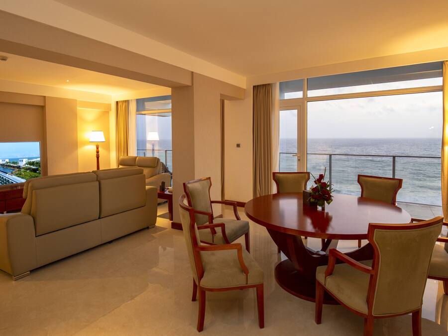 A hotel room with a large TV, a sofa, a dining table with chairs, and a view of the ocean.