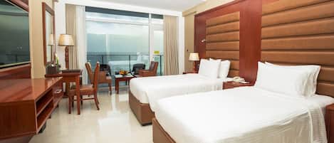 A hotel room with two beds, a desk, a chair, a small table with a vase, and a view of the sea.