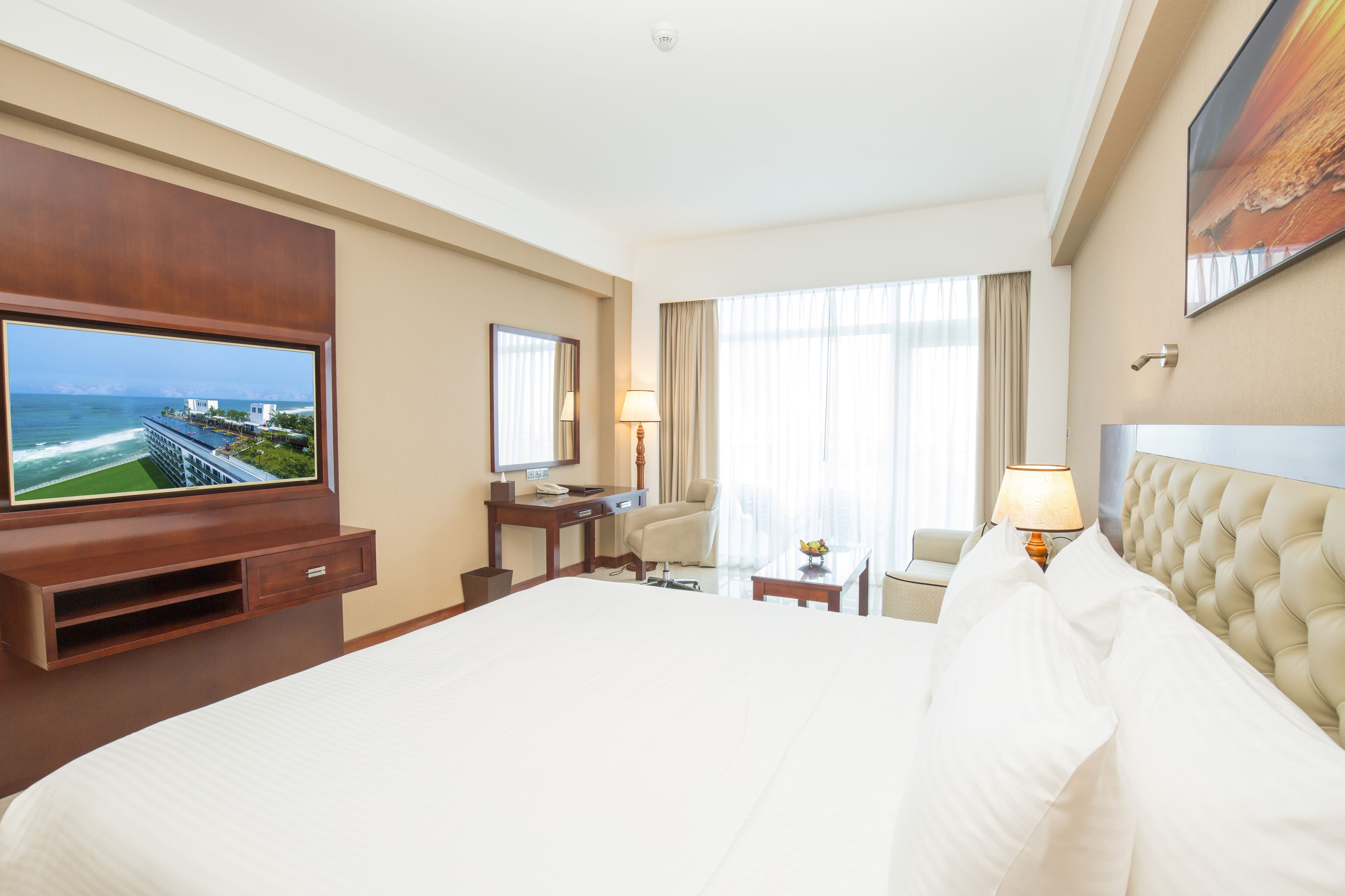 Een hotelkamer met een groot bed, een aan de muur bevestigde televisie, een bureau met stoel, een lamp en uitzicht op de oceaan door een raam.
