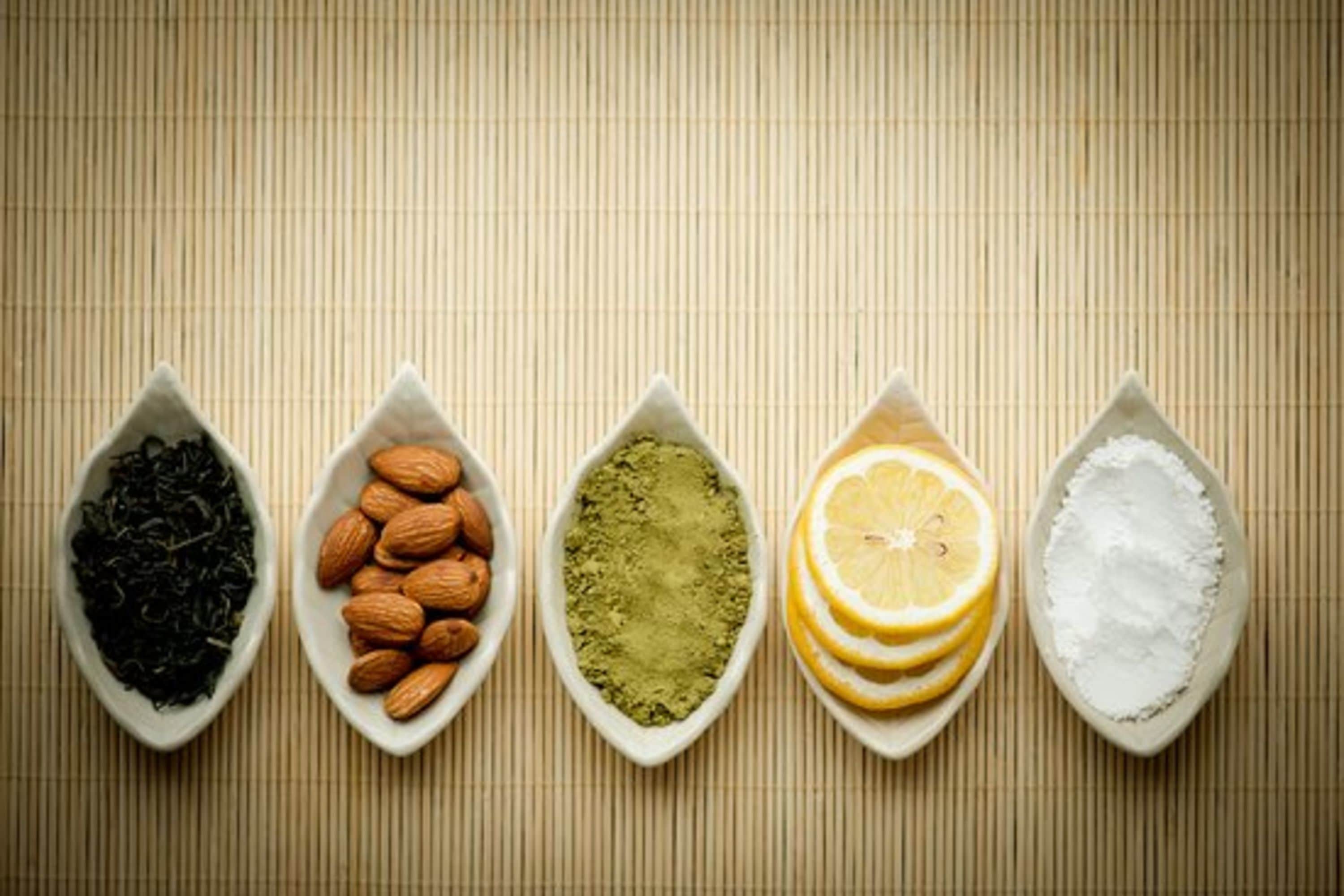 Five white ceramic spoons containing different items: black tea leaves, almonds, green matcha powder, lemon slices, and white powder.