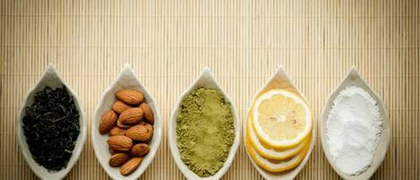 Five white ceramic spoons containing different items: black tea leaves, almonds, green matcha powder, lemon slices, and white powder.