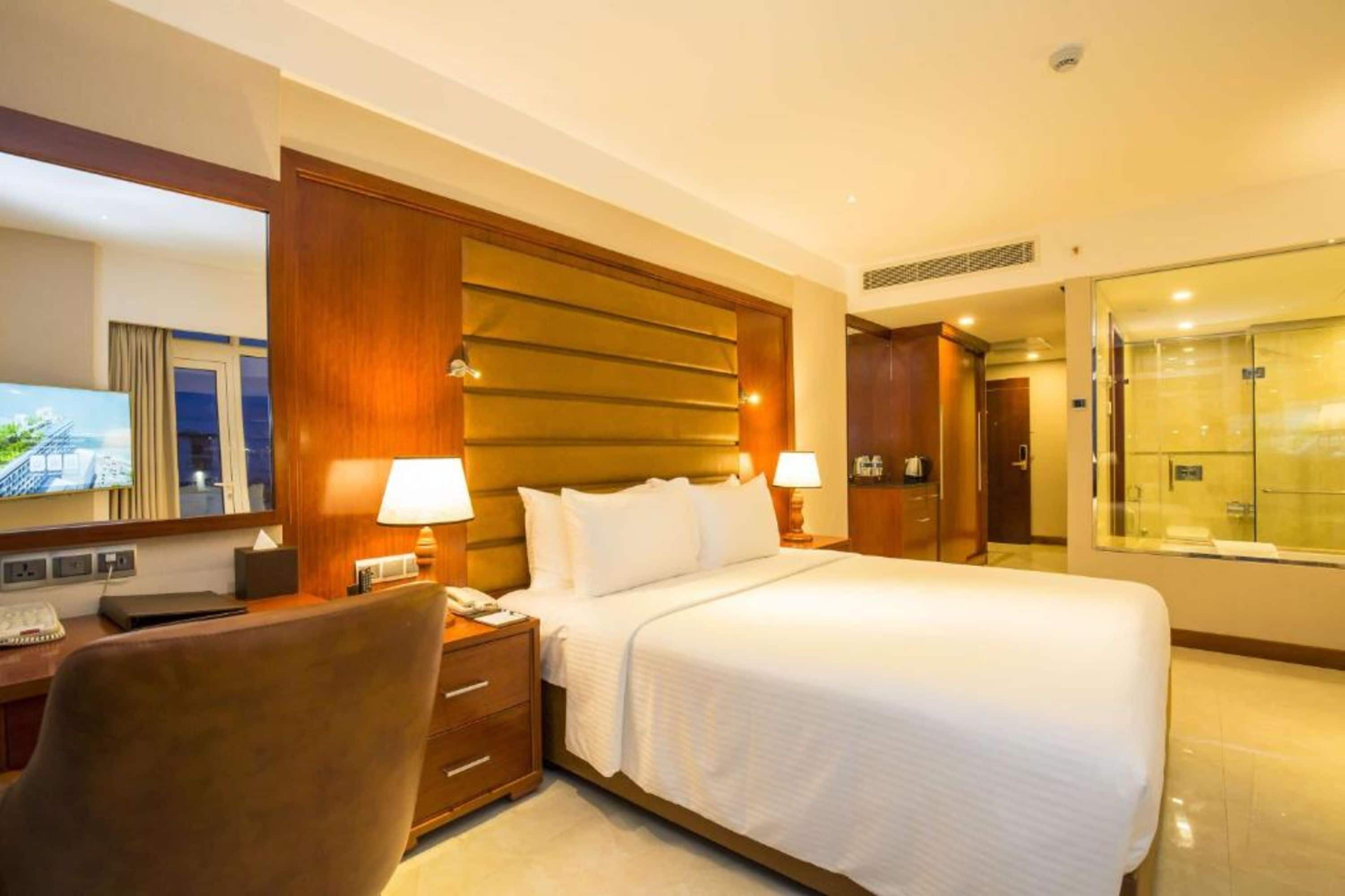 A hotel room with a large bed, two bedside tables with lamps, a desk with a computer, a chair, a TV, and a glass shower area.