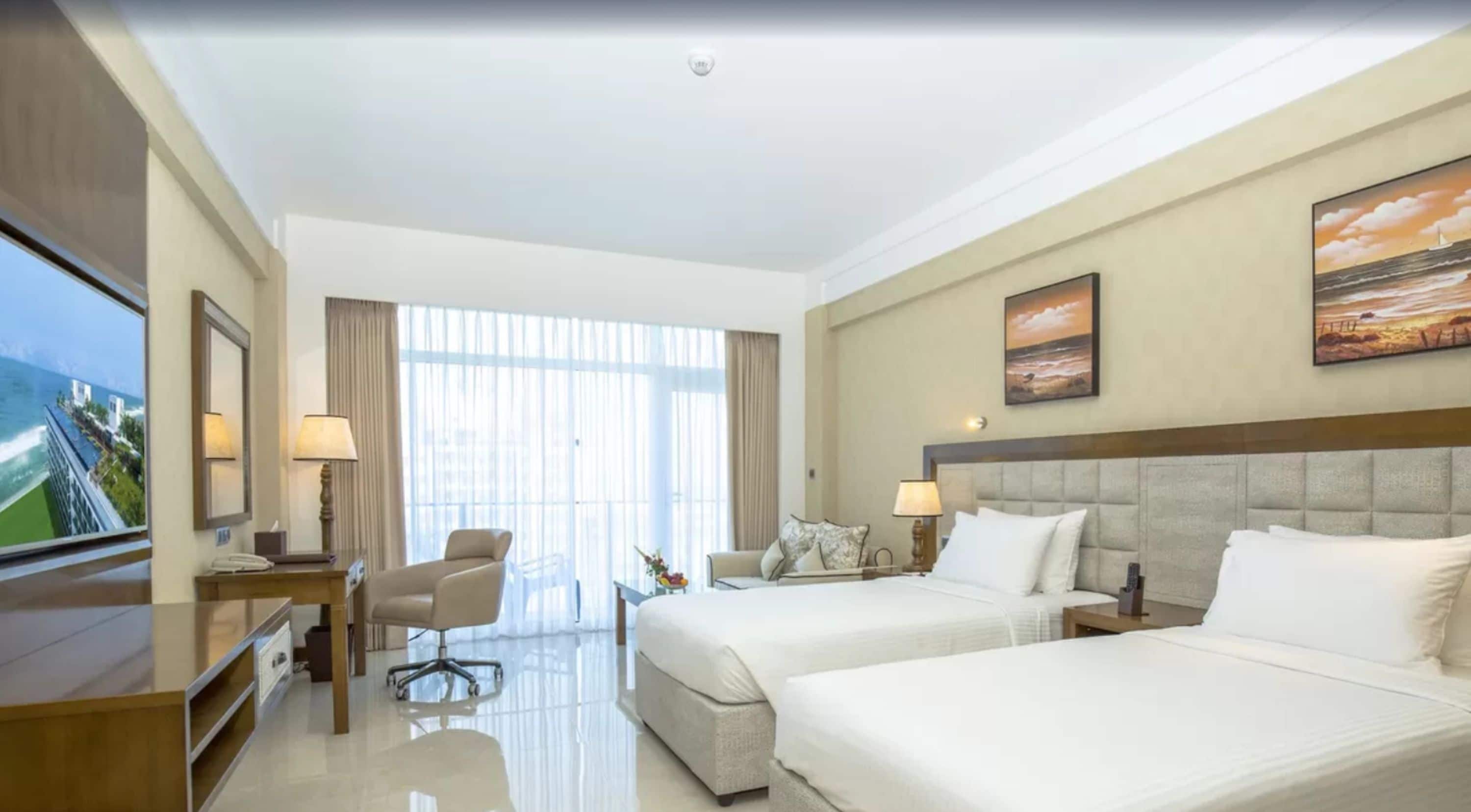 A hotel room with two beds, a desk, a chair, and a large window with a view of the ocean.