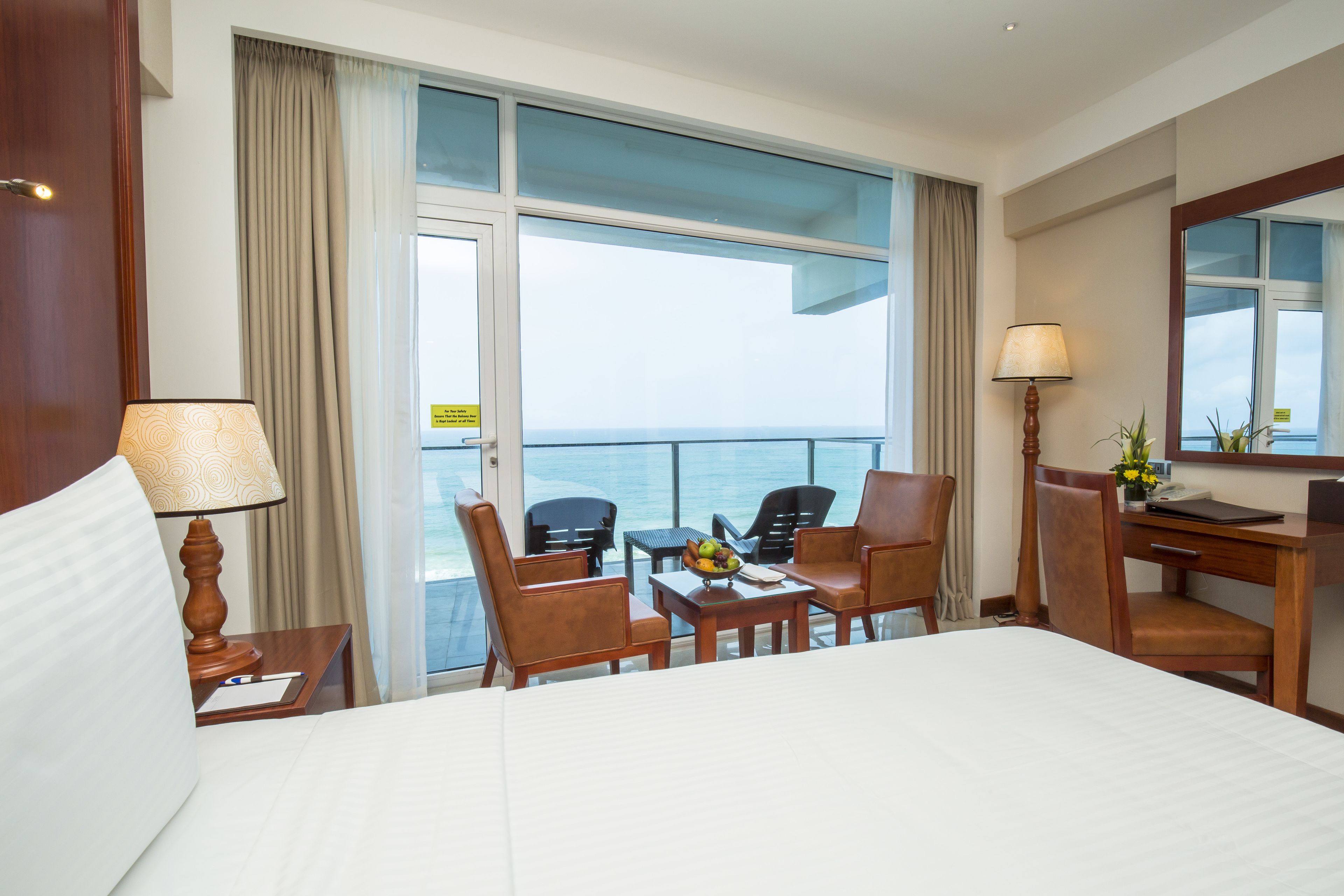 Ein Hotelzimmer mit einem Bett, einem Schreibtisch, zwei Stühlen, einem Tisch mit Obstschale, einer Lampe und Meerblick durch große Fenster.