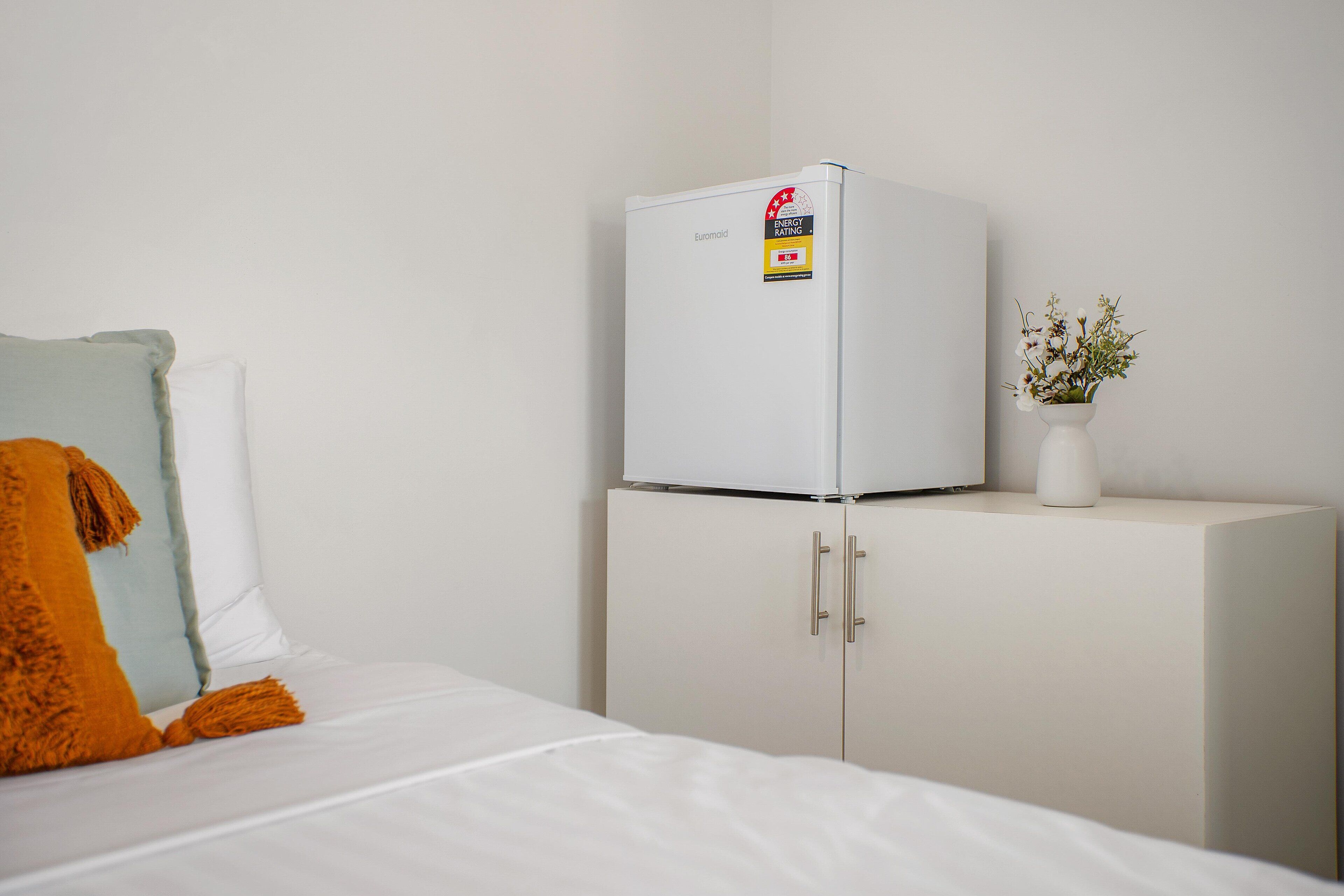 A white bed with an orange throw blanket, a white cabinet with a refrigerator on top, and a vase with flowers on the cabinet.
