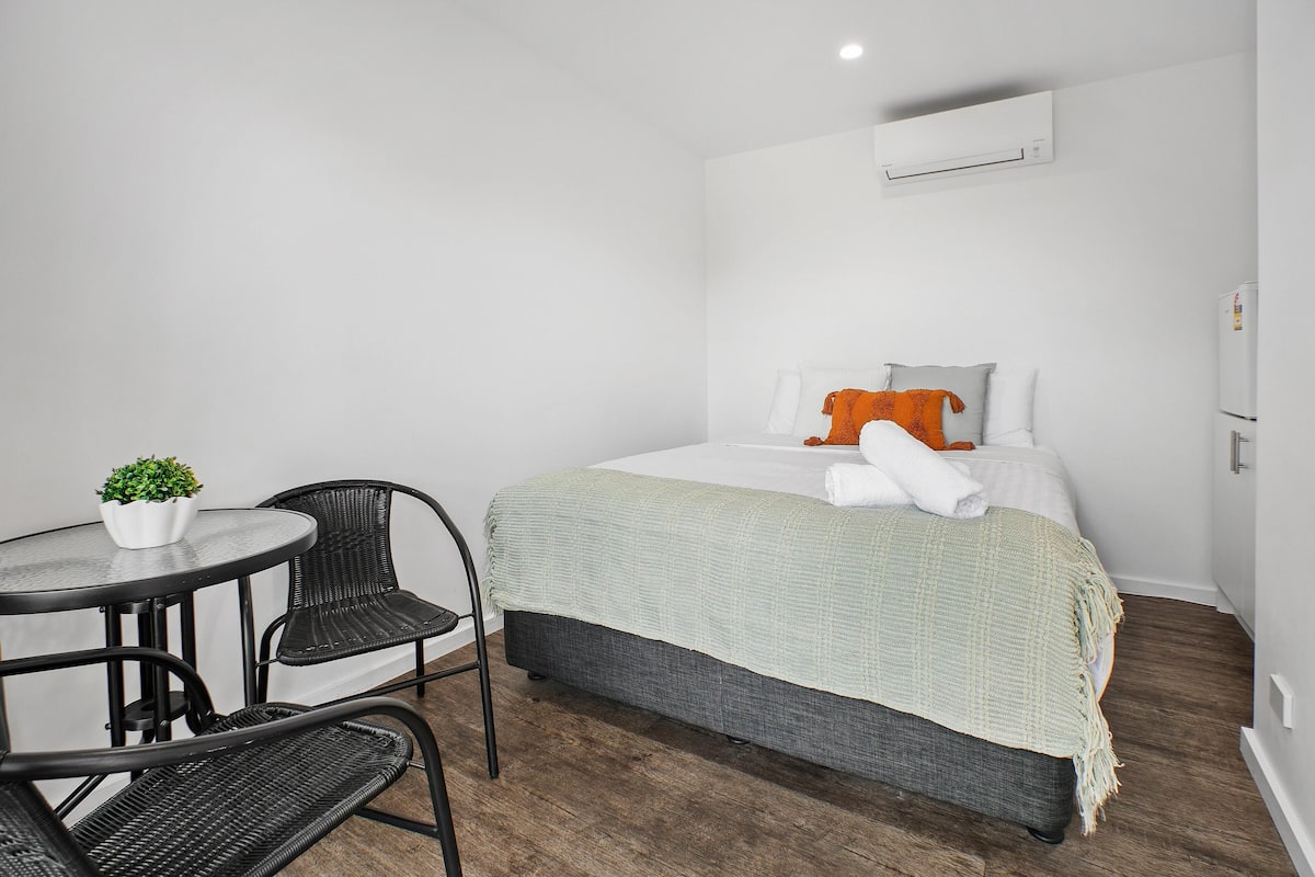 A bedroom with a bed, a small table with chairs, a potted plant, and an air conditioning unit.