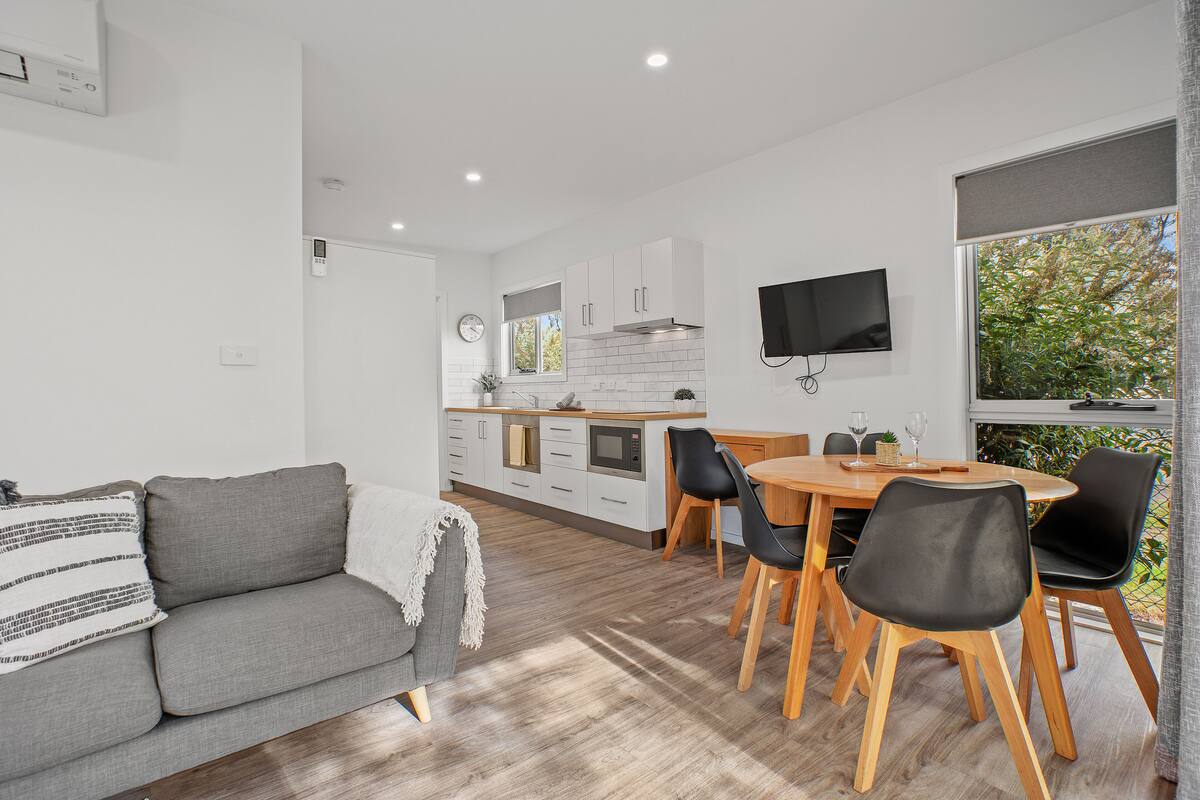 A modern kitchen with white cabinets, a dining area with a wooden table and black chairs, and a grey sofa with a throw blanket.