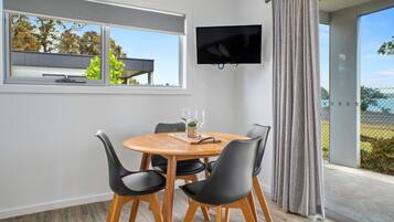 Een eethoek met een ronde houten tafel, zwarte stoelen, een aan de muur bevestigde televisie en uitzicht op het buiten via een raam.