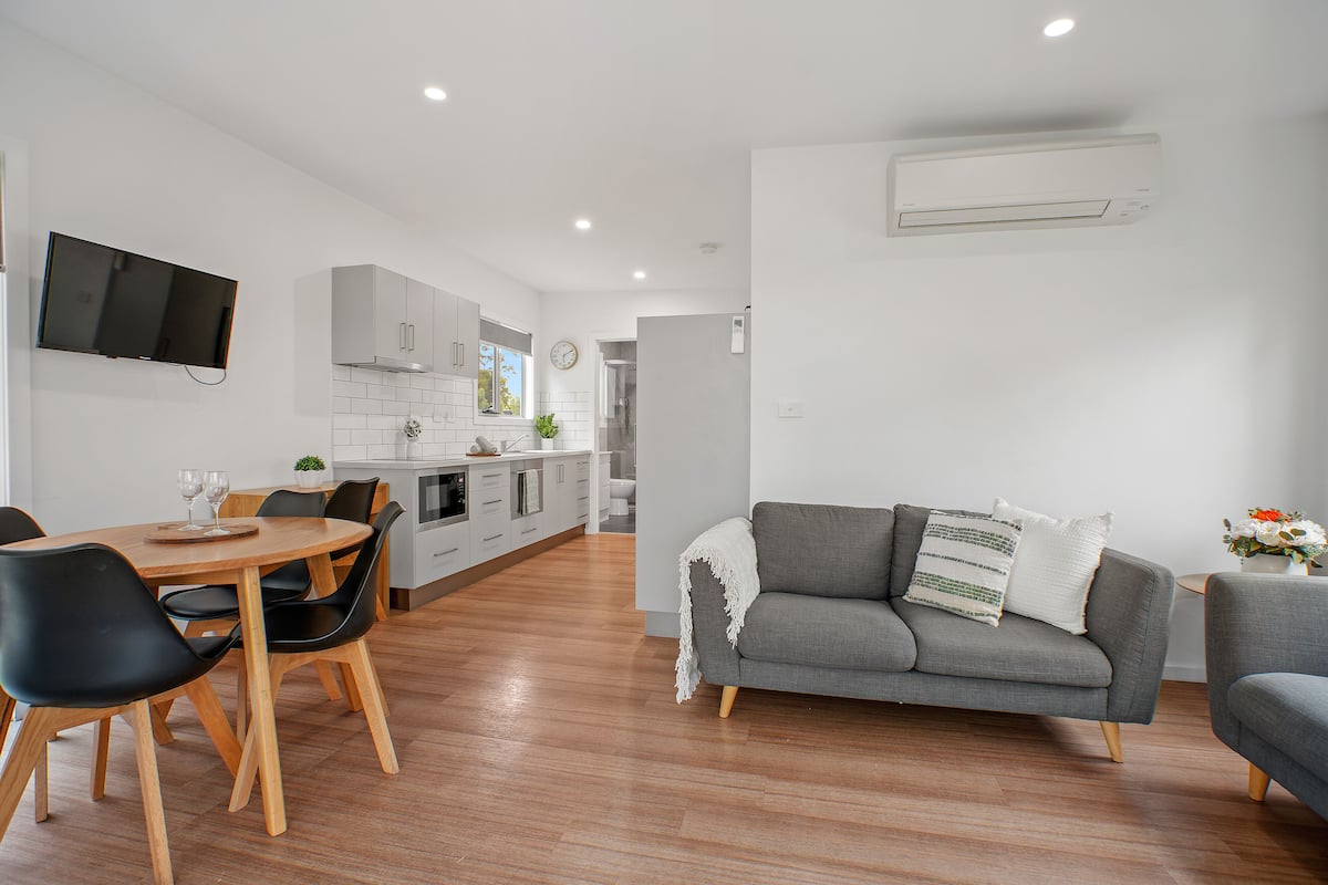 A modern living room with a grey sofa, a dining table with chairs, and a kitchen in the background.