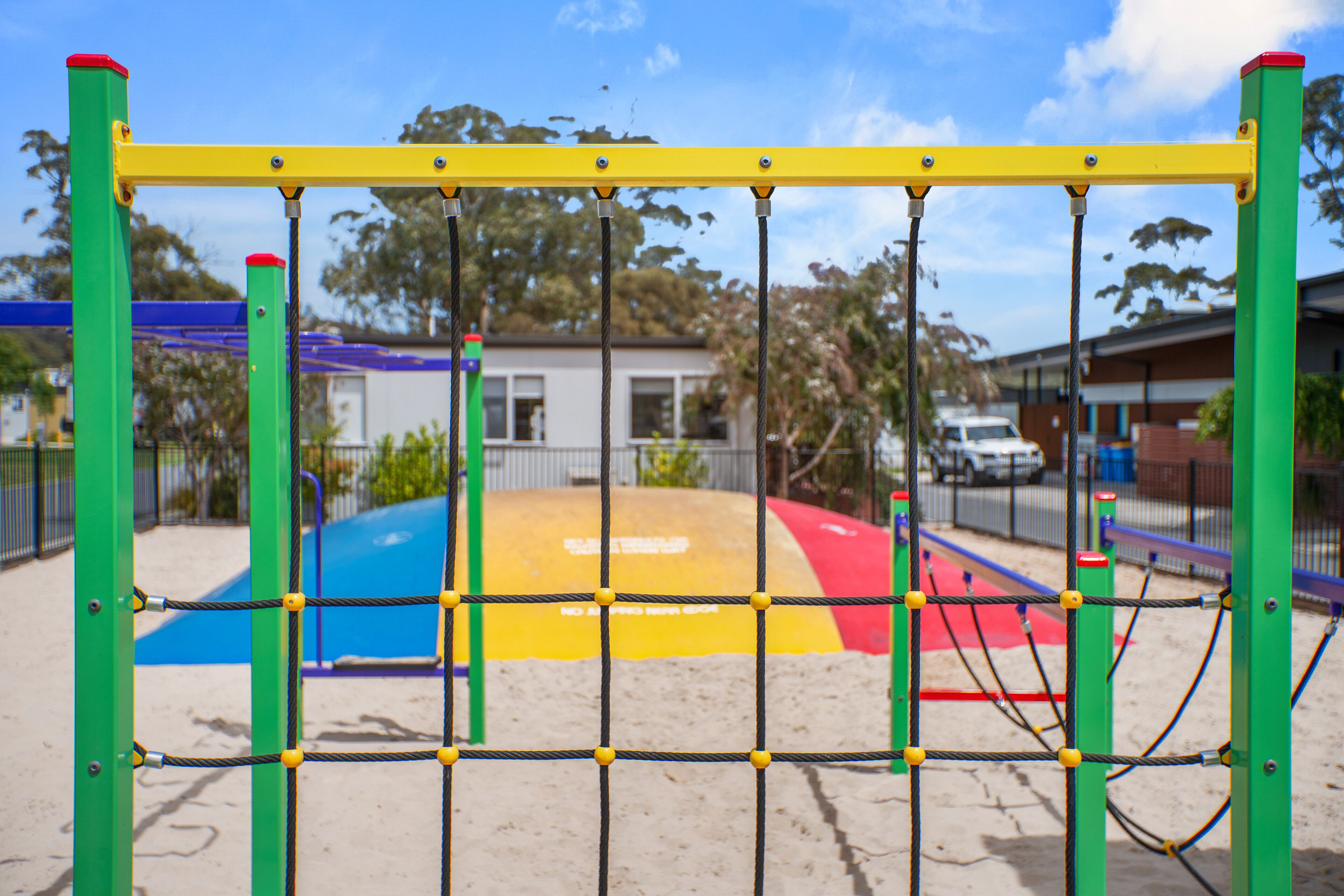 A playground with a yellow and green climbing structure, a blue and red slide, and a fenced area.
