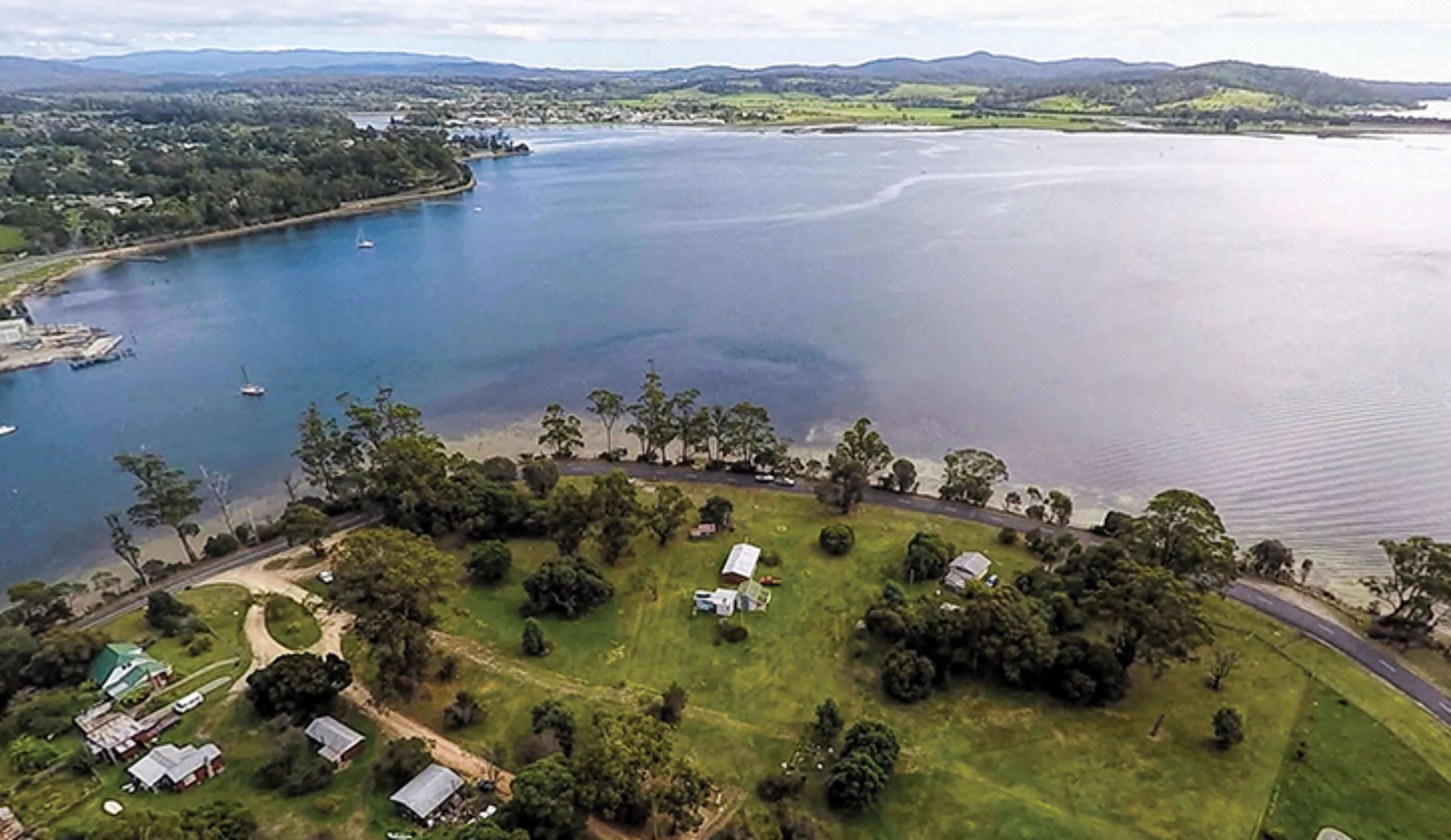 Vue aérienne d’une zone côtière comprenant un port de plaisance, des champs verdoyants et quelques bâtiments dispersés.