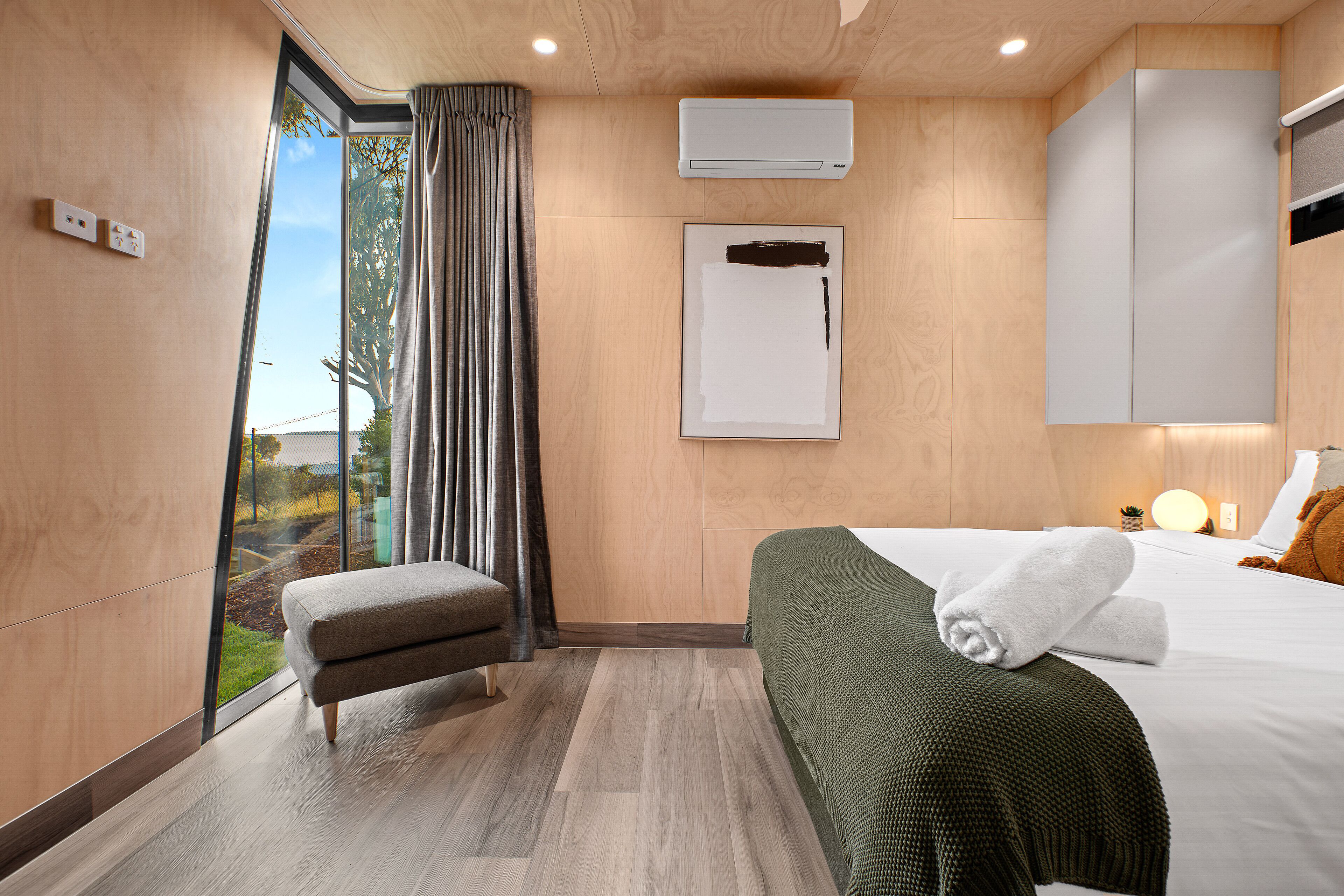 A modern hotel room with a large bed, a bench, a view of the outdoors, and an air conditioning unit.