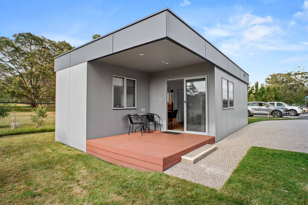 A modern single-story house with a flat roof, a covered patio with a table and chairs, and a driveway with parked cars.