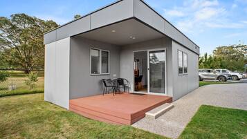 Una casa moderna de un piso con techo plano, un porche cubierto con una mesa y sillas, y un camino de entrada con coches aparcados.