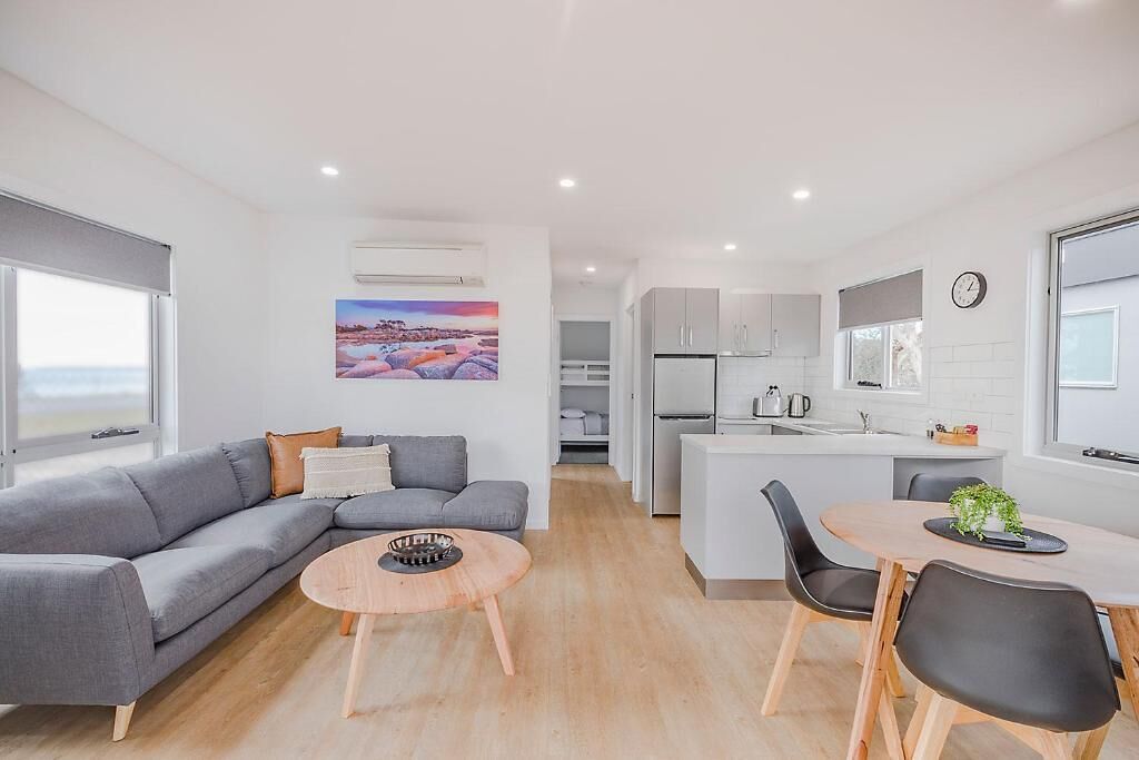 Een moderne woonkamer met een grijze bank, een ronde houten salontafel, en een eethoek met een kleine tafel en stoelen. In de achtergrond is een keuken met witte kastjes en een spoelbak zichtbaar.