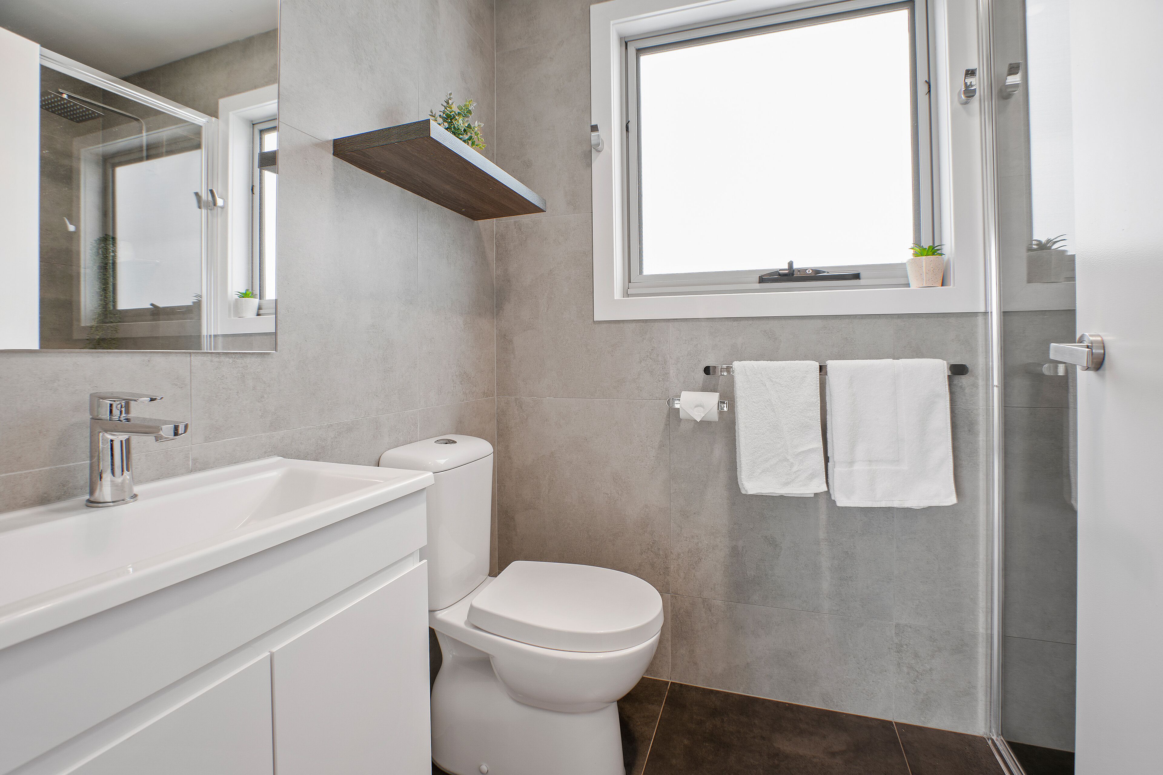 A modern bathroom with a white sink, toilet, and shower. There is a window above the toilet and a shelf with a plant.