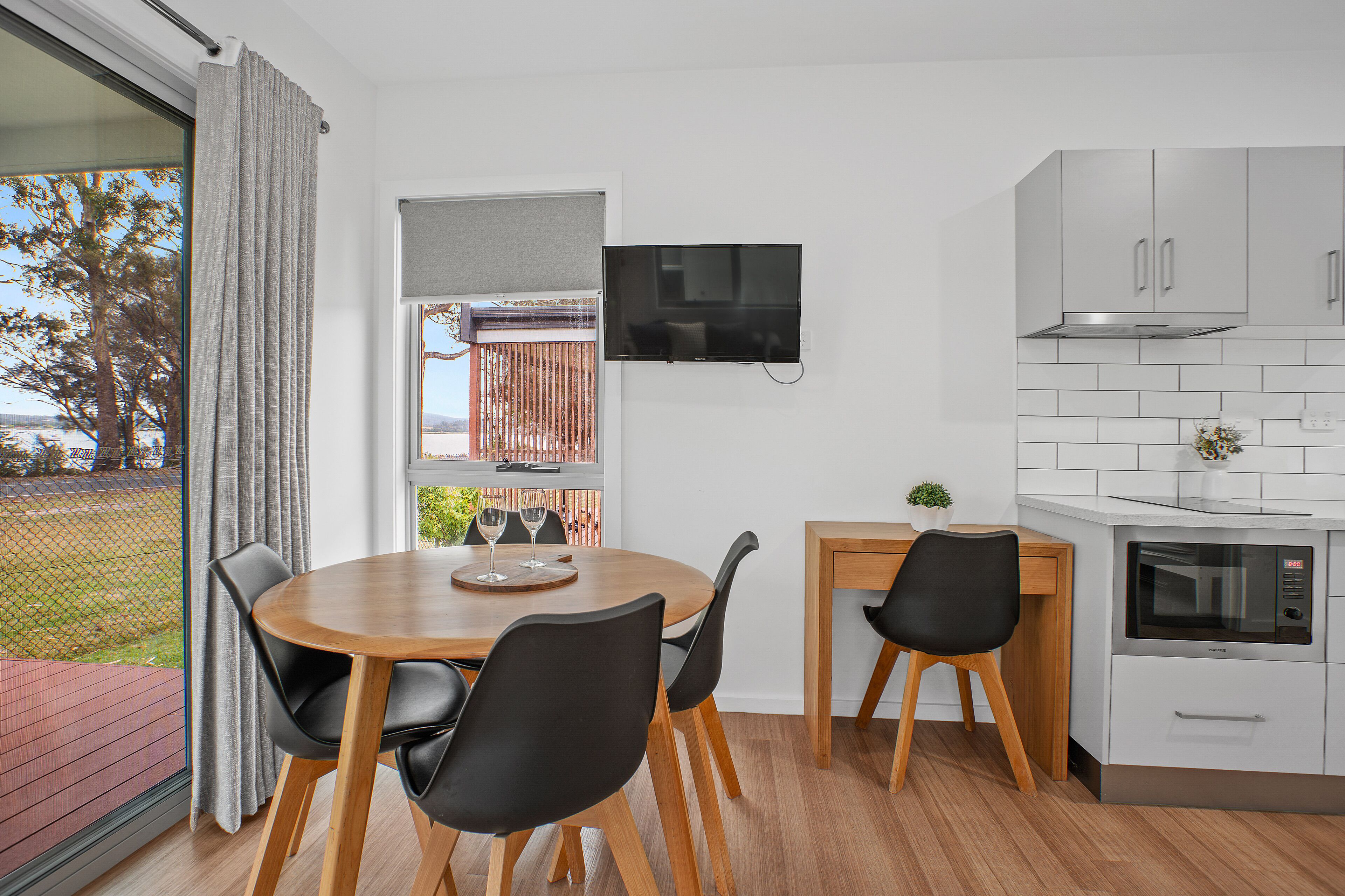 A modern kitchen with a dining area, a TV mounted above the counter, and a view of the outdoors through a window.