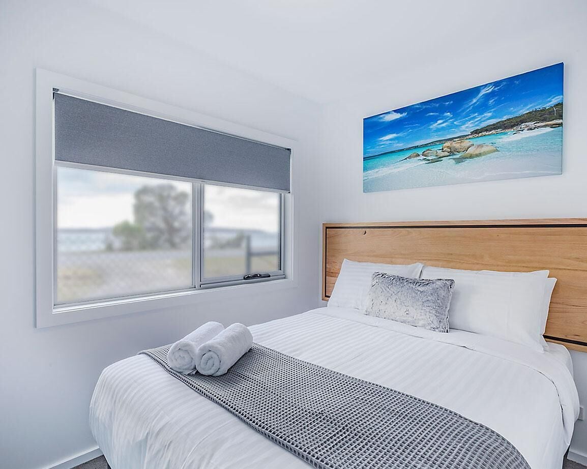 A bedroom with a large window, a bed with white bedding, a wooden headboard, and a painting of a beach scene.