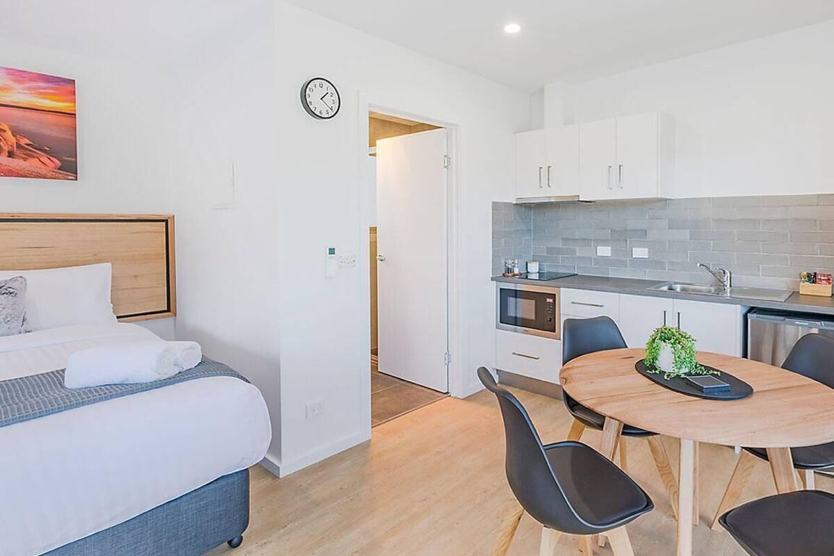 A modern hotel room with a bed, a dining table with chairs, a kitchen area, and a clock on the wall.