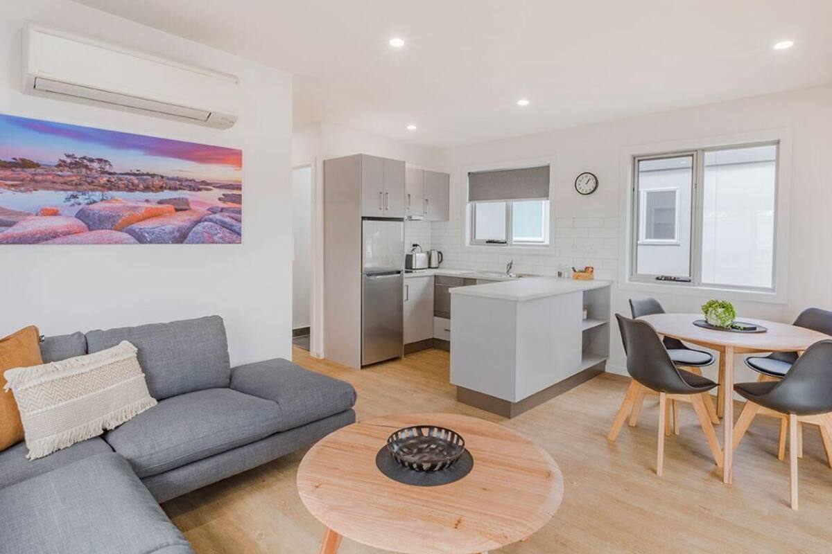 A modern apartment with a grey sofa, a round wooden coffee table, a kitchen with white cabinets, and a dining area with a table and chairs.