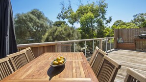 Eine Holzterrasse mit Tisch und Stühlen, einer Obstschale und Blick auf Bäume und einen Zaun.