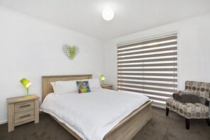 Un dormitorio con cama de madera, ropa de cama blanca, una lámpara verde, una decoración con forma de corazón y una silla con tapicería estampada.