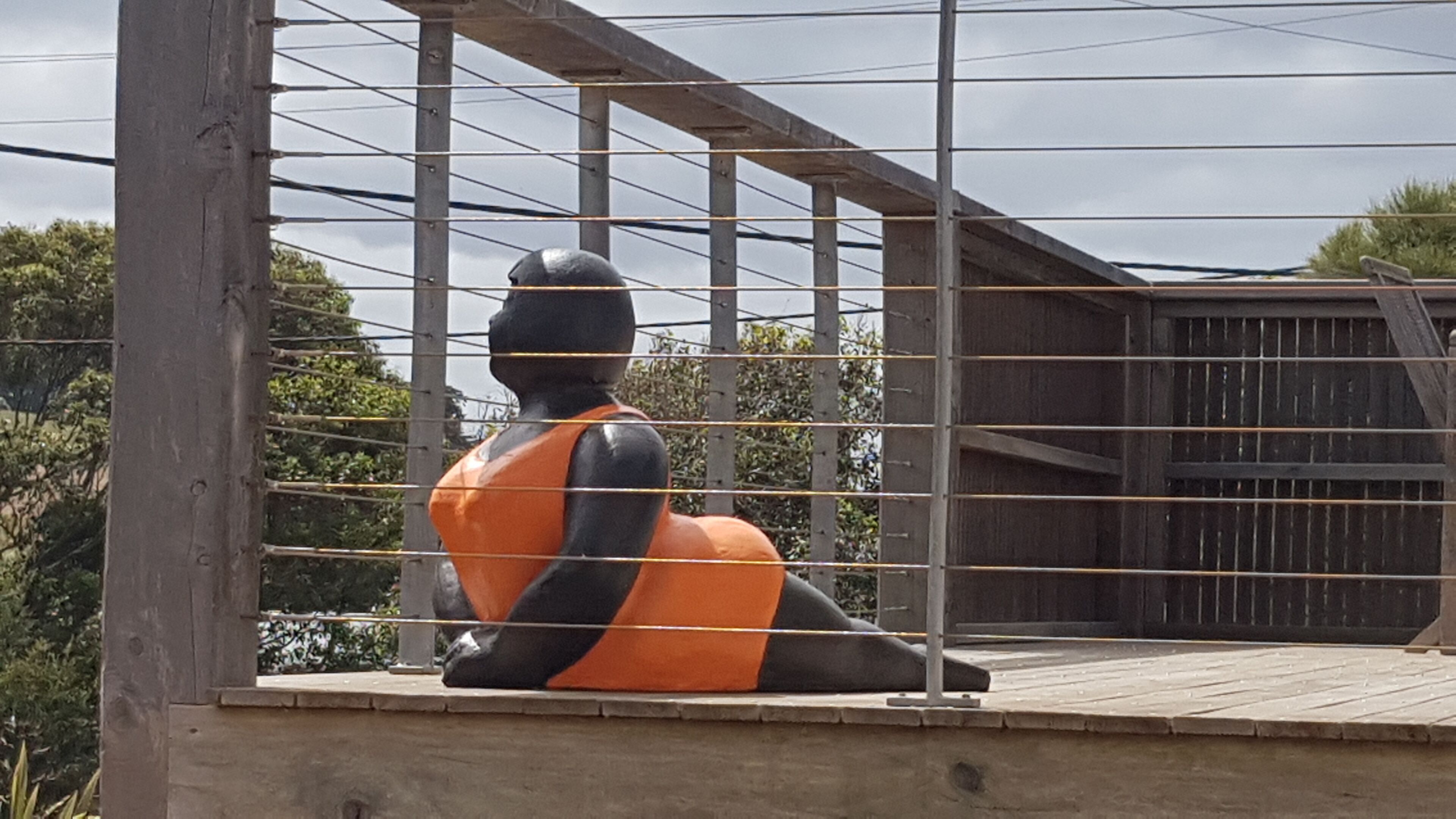 Une sculpture représentant une figure assise, vêtue d’une tenue orange et noire, et entourée d’une clôture métallique.