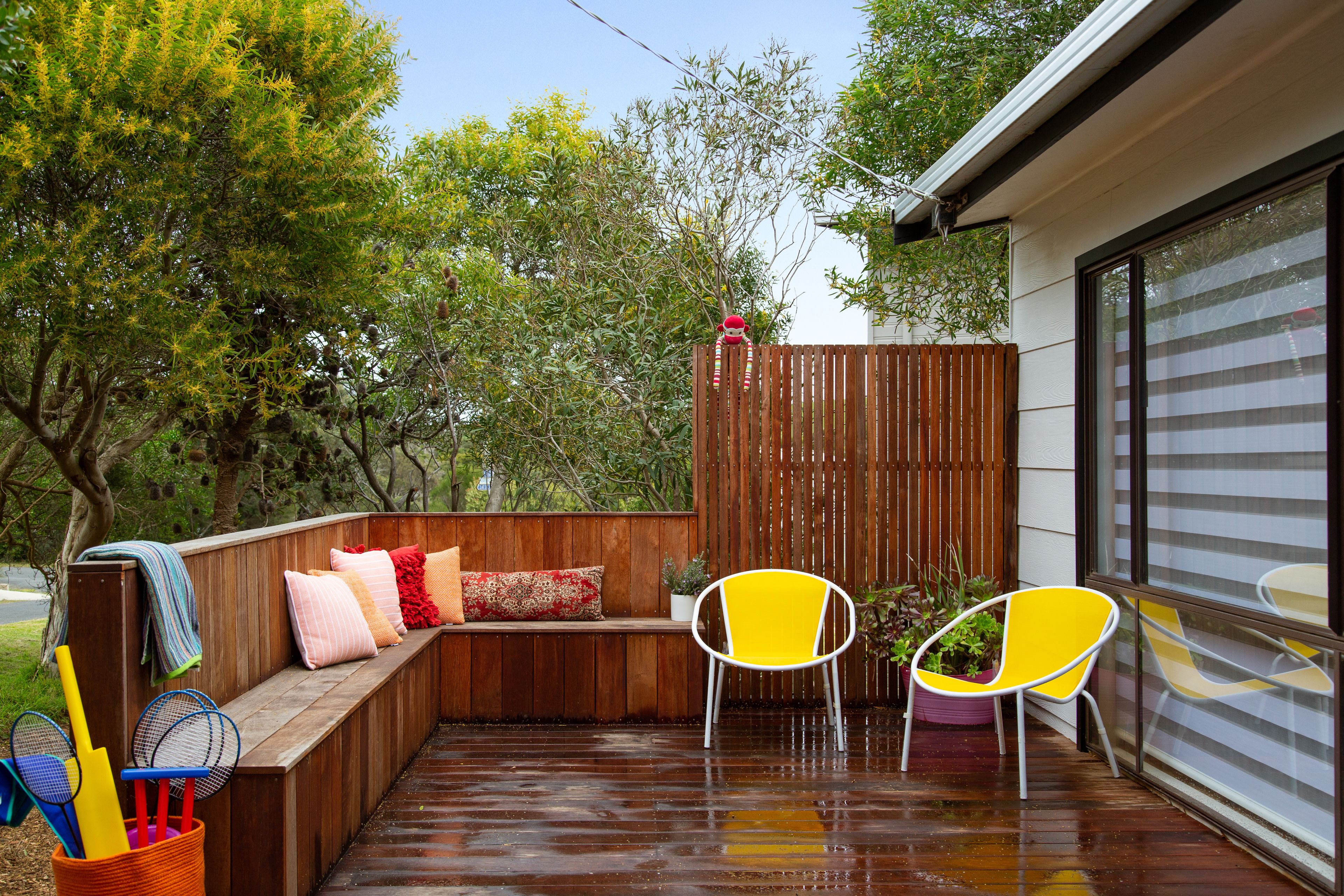 Une terrasse en bois avec un banc et des chaises, entourée d’arbres et d’une clôture.