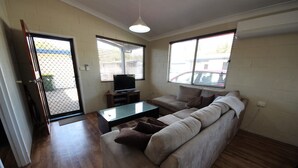 Una sala de estar con un sofá, una mesa de centro y un televisor. Hay ventanas con persianas y una puerta que da al exterior.