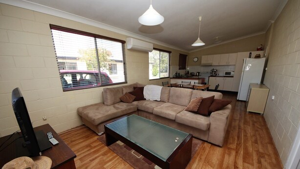 Una sala de estar con un sofá, una mesa de centro de vidrio y un televisor. Se ve una cocina al fondo con un refrigerador y un microondas.