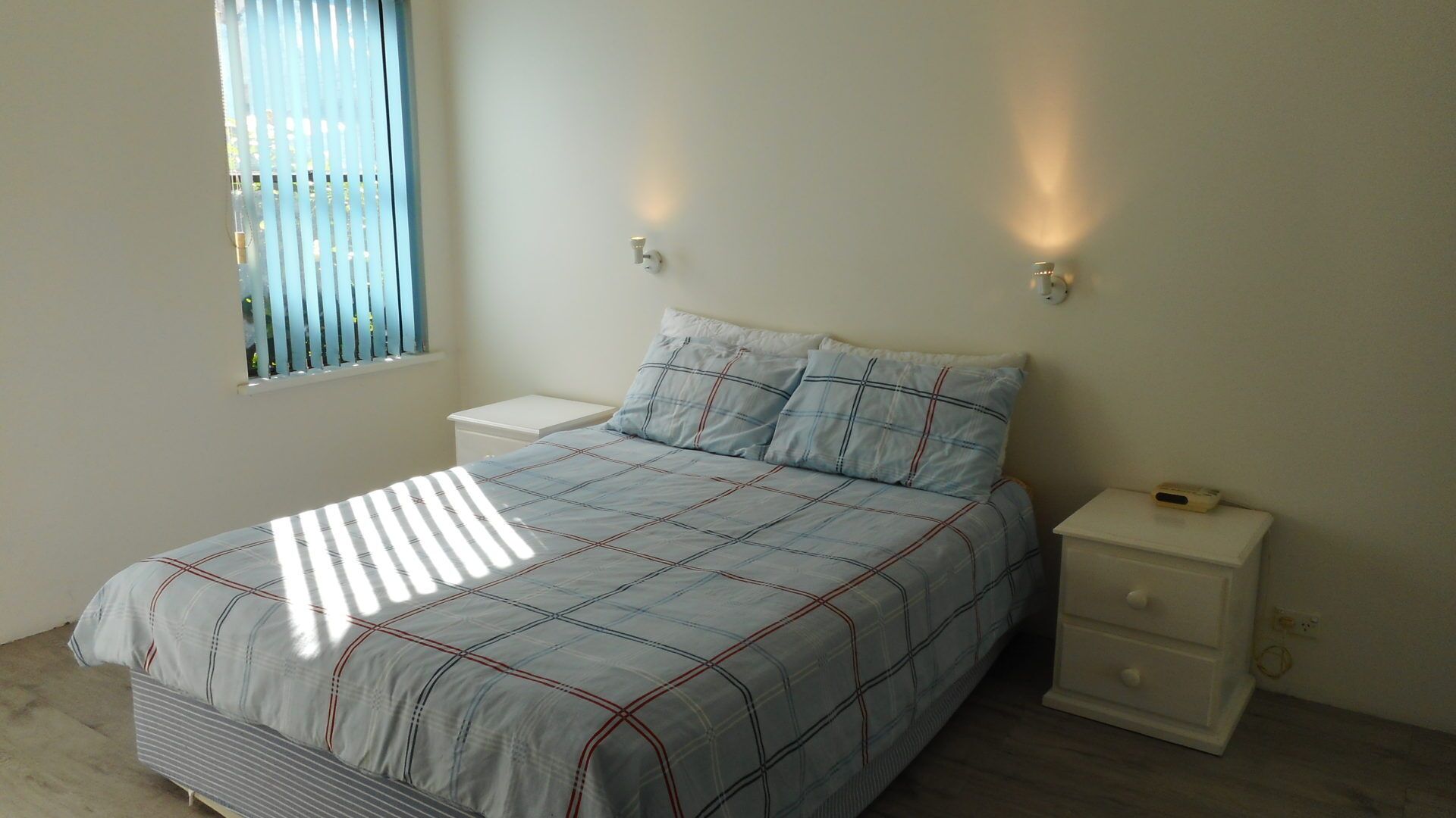 Une chambre à coucher comprenant un lit, deux tables de chevet, une fenêtre avec des stores et des lampes fixées au mur.