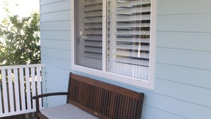 Um banco de madeira num deck de madeira com guarda-corpo branco, uma janela com persianas e uma parede exterior azul-claro.