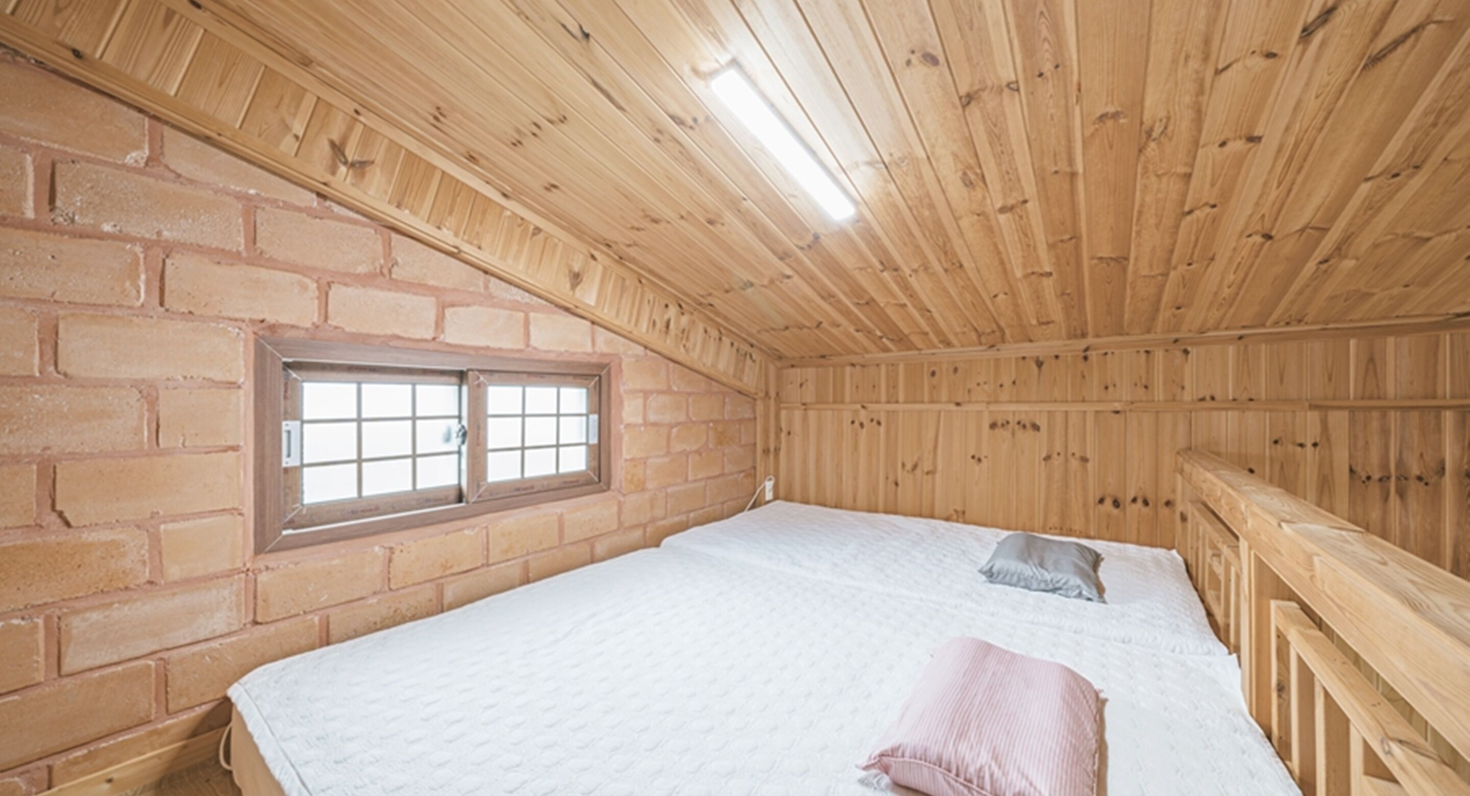 Une pièce avec un plafond en bois, des murs en briques, un lit simple avec des oreillers et une fenêtre avec des barreaux.