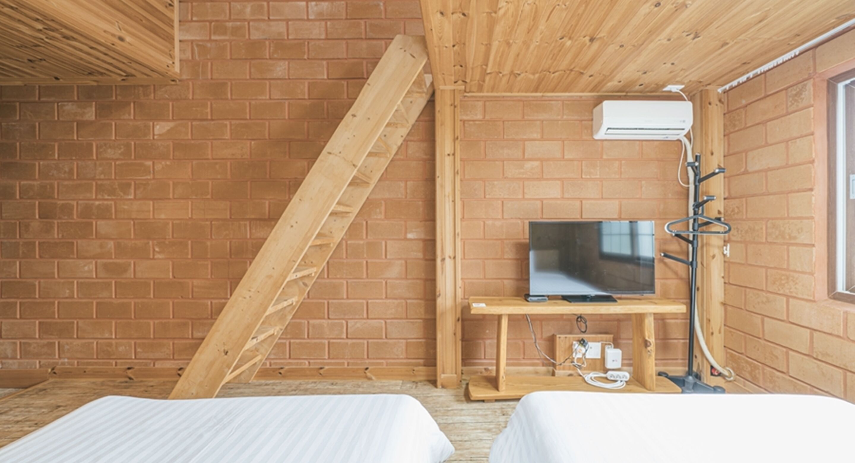 Ein Zimmer mit Backsteinwand, schrägverdeckter Holzbalkendecke, einem Fernseher auf einem Holzständer und einem Fenster.